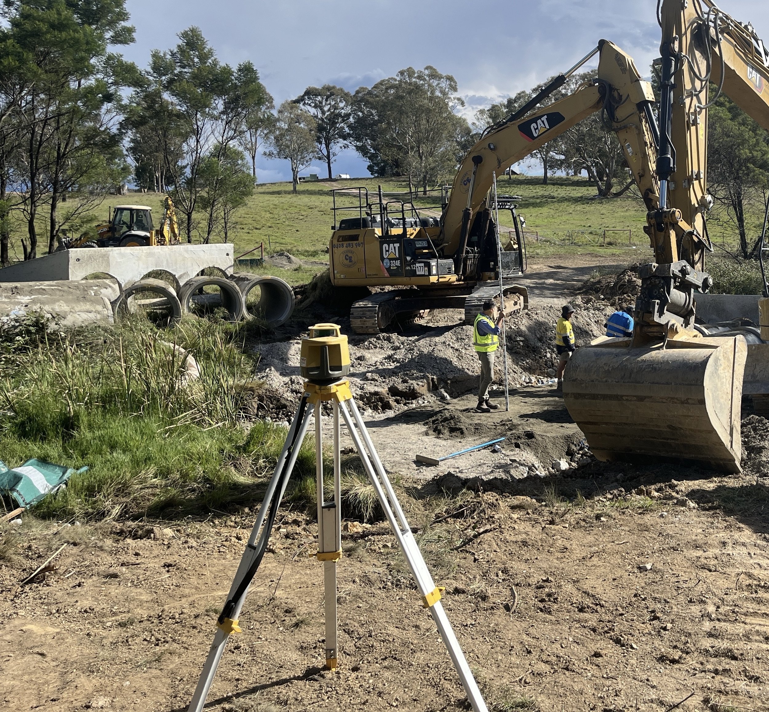 Construction site with a large yellow excavator and workers wearing safety vests, surrounded by trees and a blue sky.