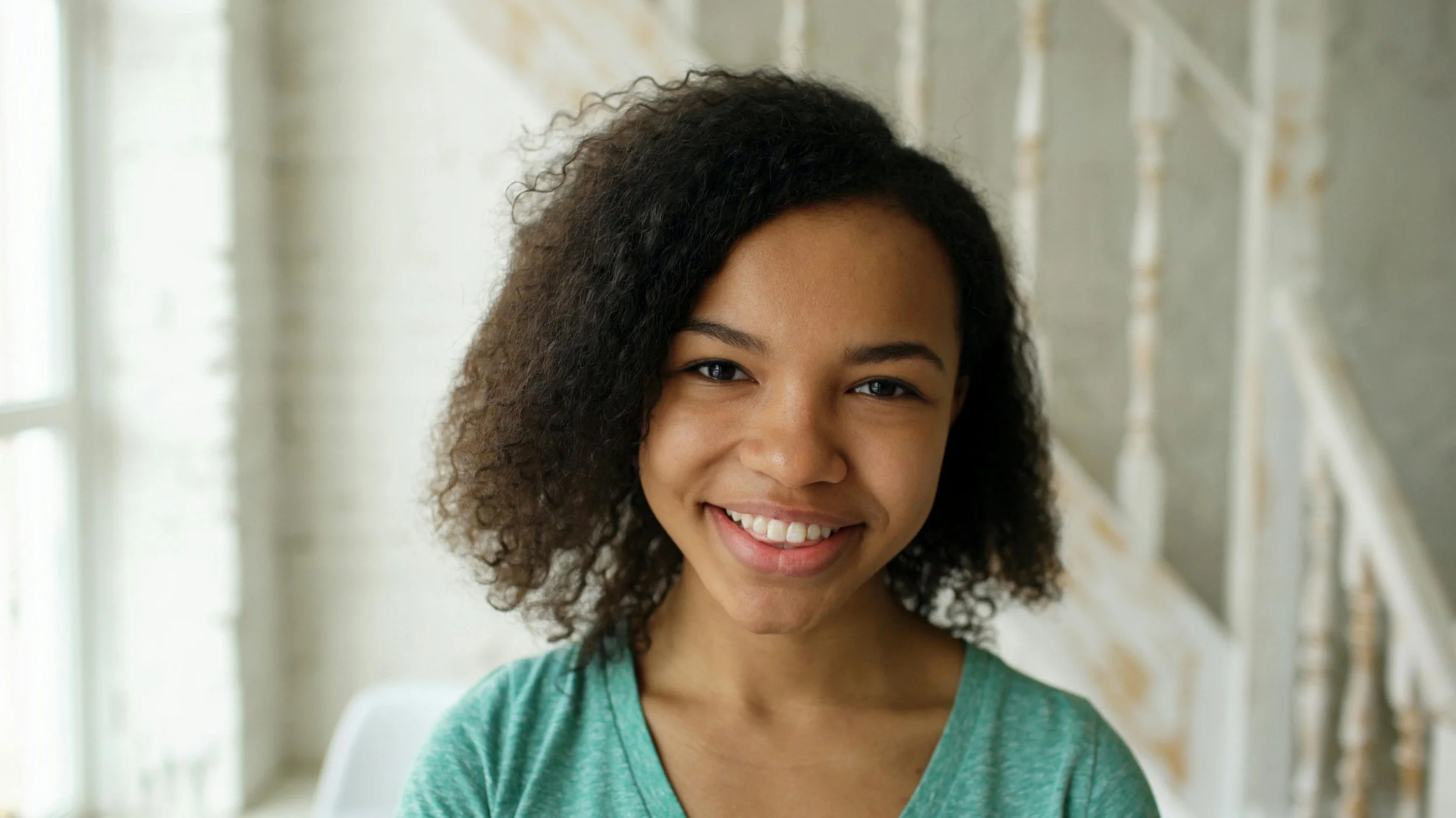 A young woman with curly hair smiling in a bright room with large windows.