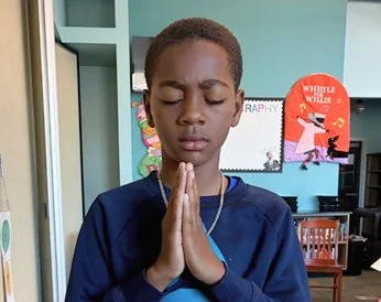 A young boy with braided hair wearing a navy blue shirt, closed eyes, and hands pressed together in a prayer pose indoors.