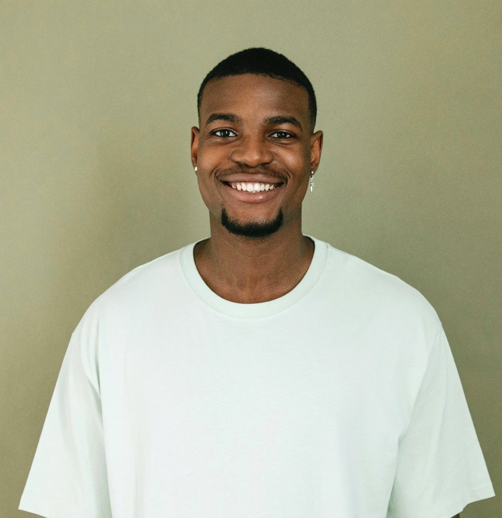 A smiling young man with wearing earrings and a white shirt, standing in front of a plain, light-colored wall.