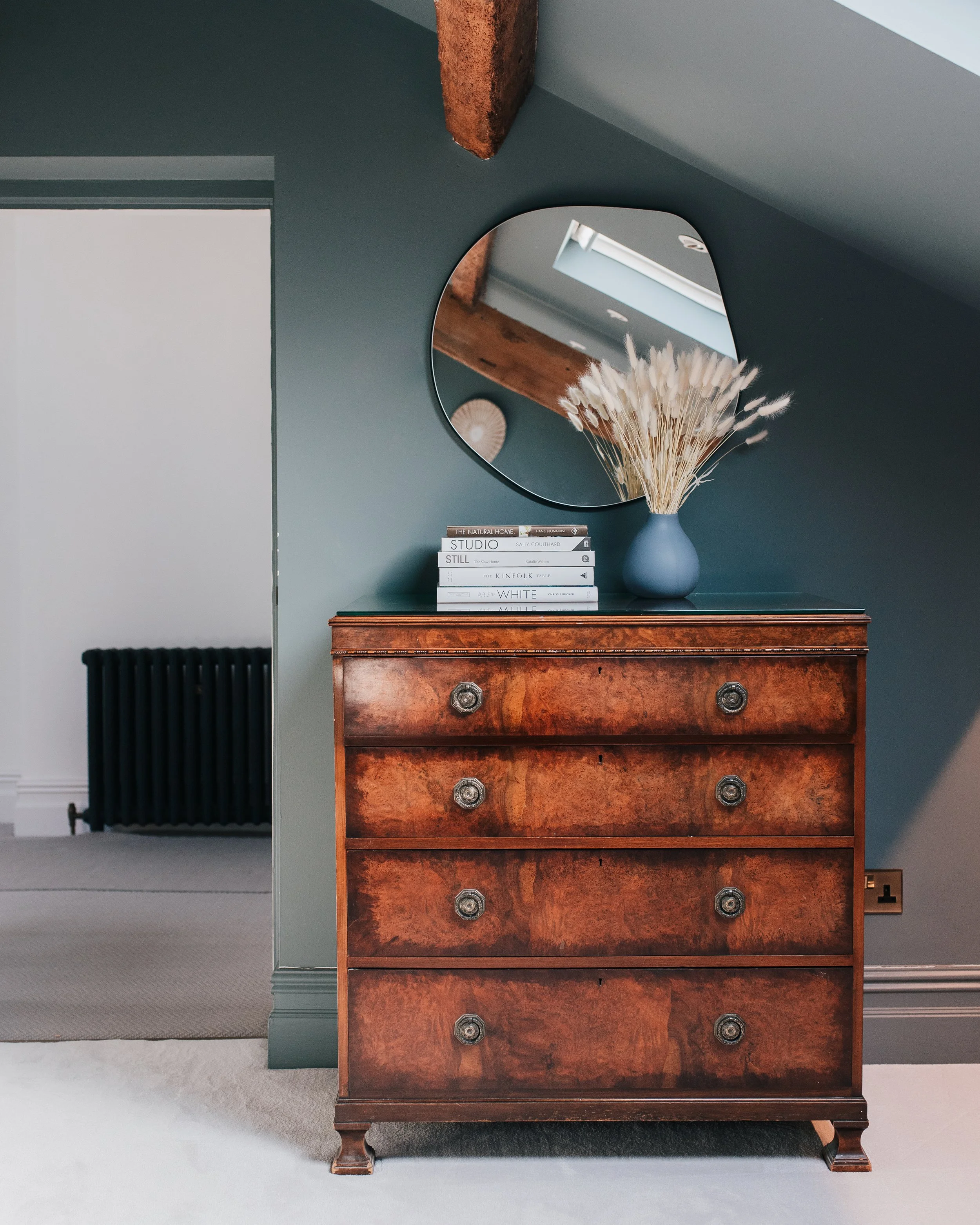 Antique wooden chest of drawers styled with books and vase beneath round mirror in bedroom