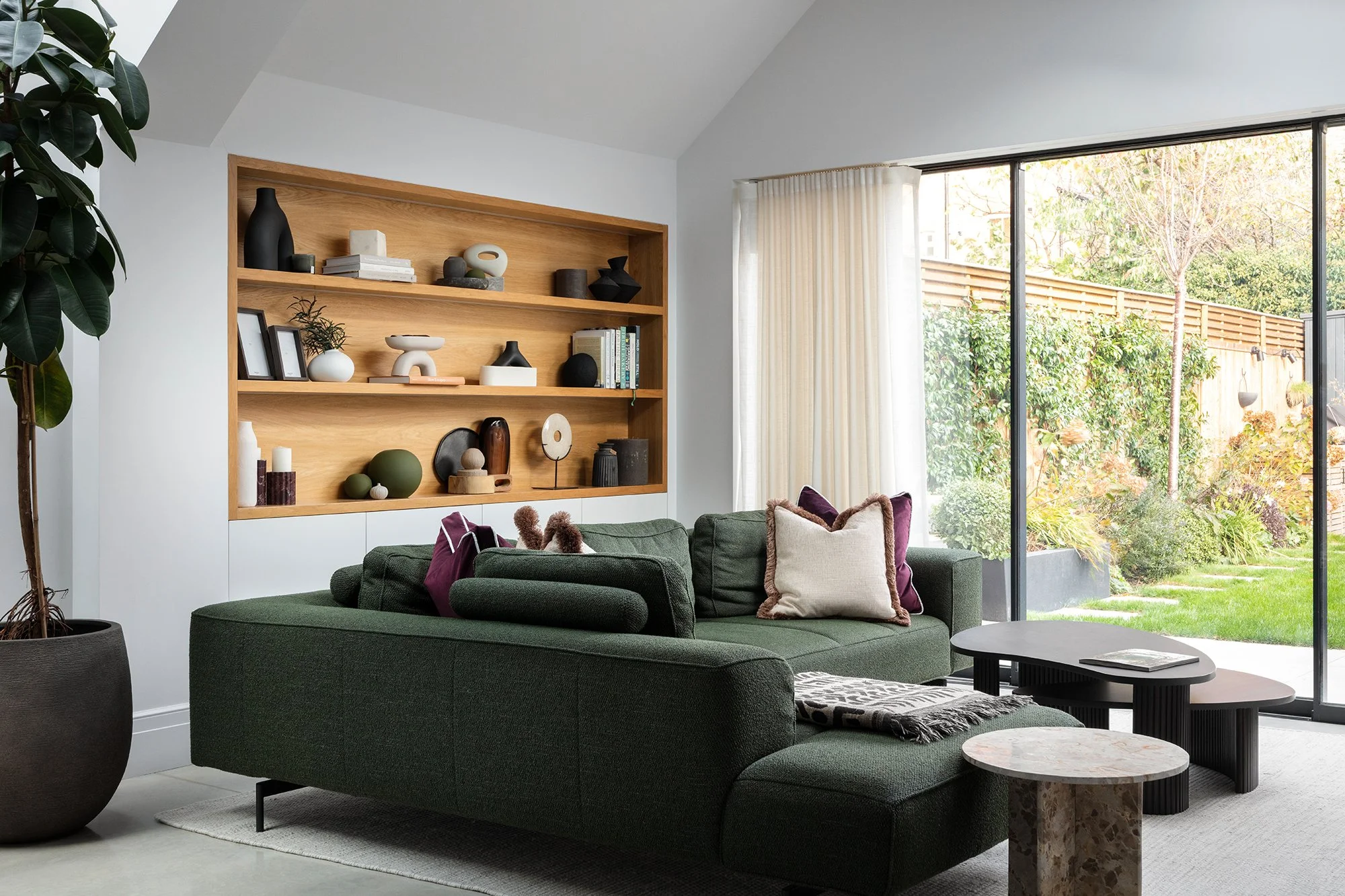 Contemporary living room with a deep green modular sofa, custom oak shelving, and curated sculptural styling designed by the studio.
