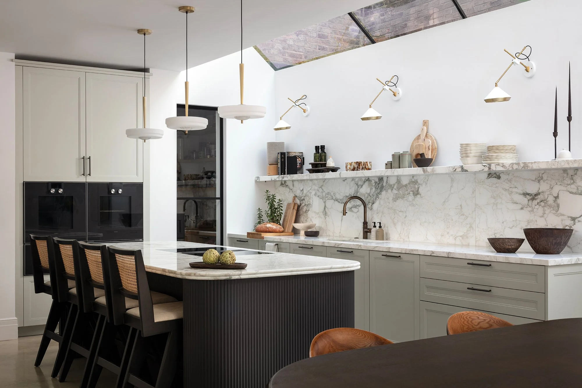 Light-filled bespoke kitchen featuring marble worktops, a fluted island, minimalist brass lighting, and curated open shelving designed by the studio.