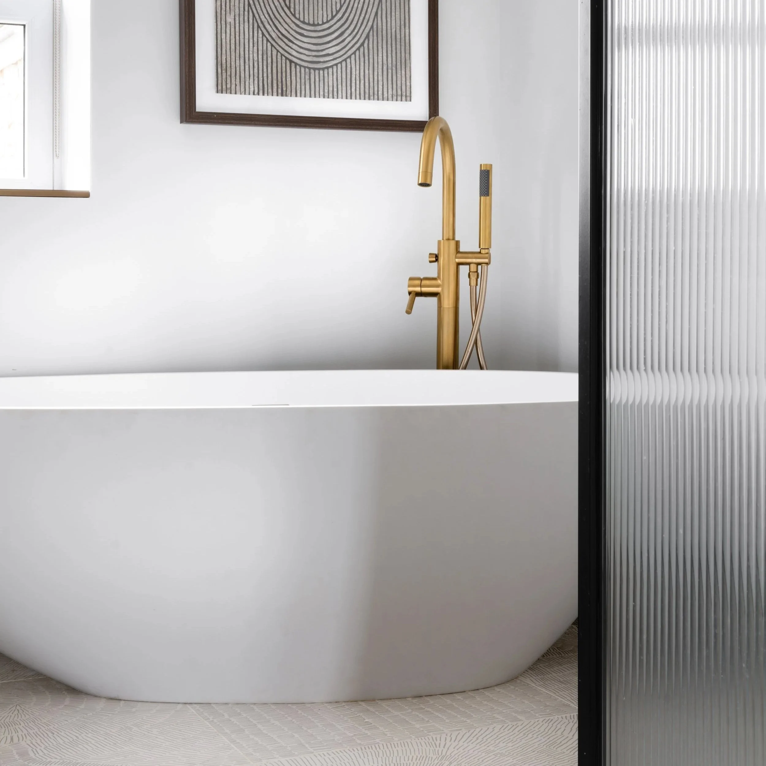 Freestanding white bath with brushed brass floor-mounted tap beside ribbed glass shower screen