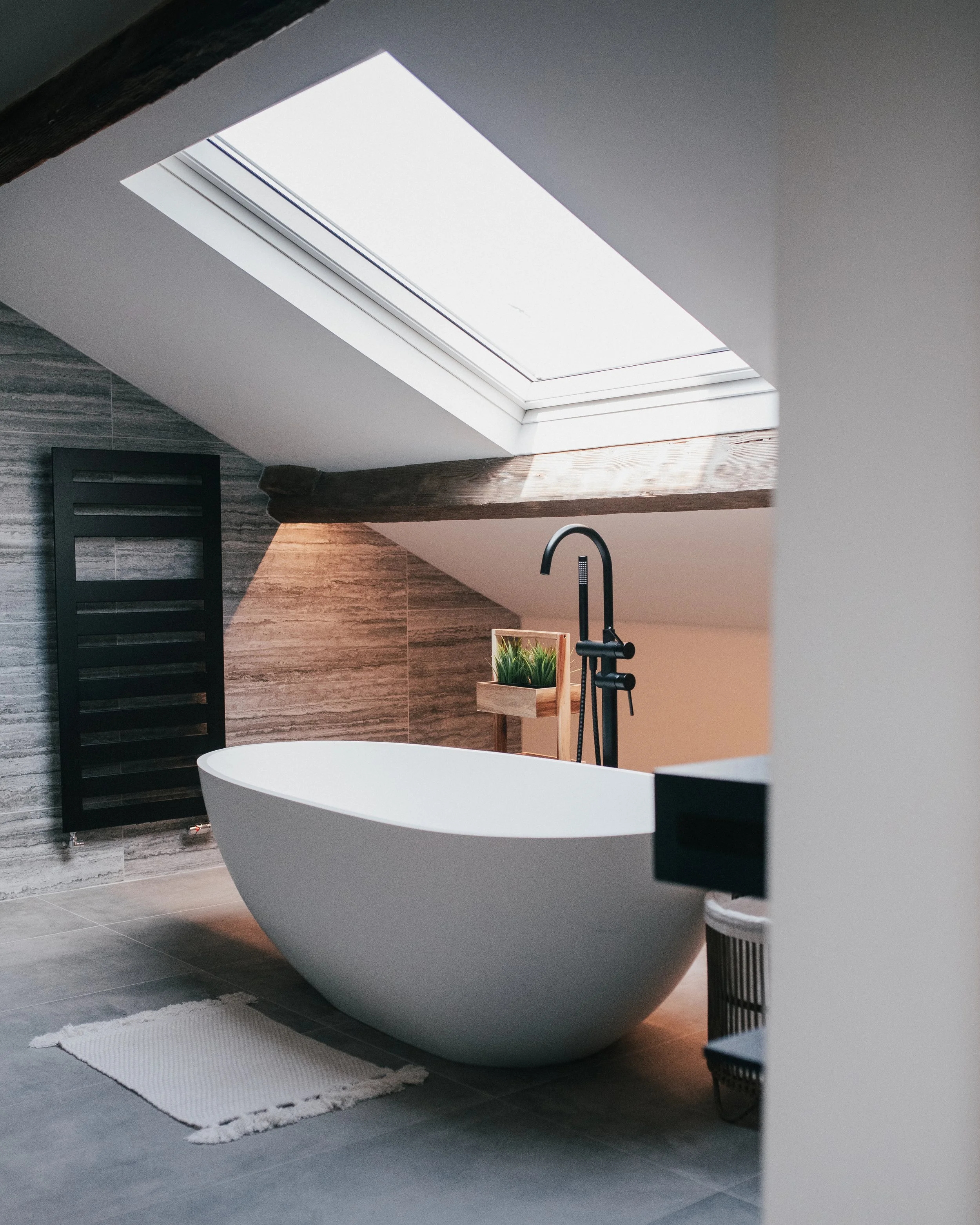 Loft bathroom with freestanding bath beneath skylight, exposed timber beam and black floor-mounted tap