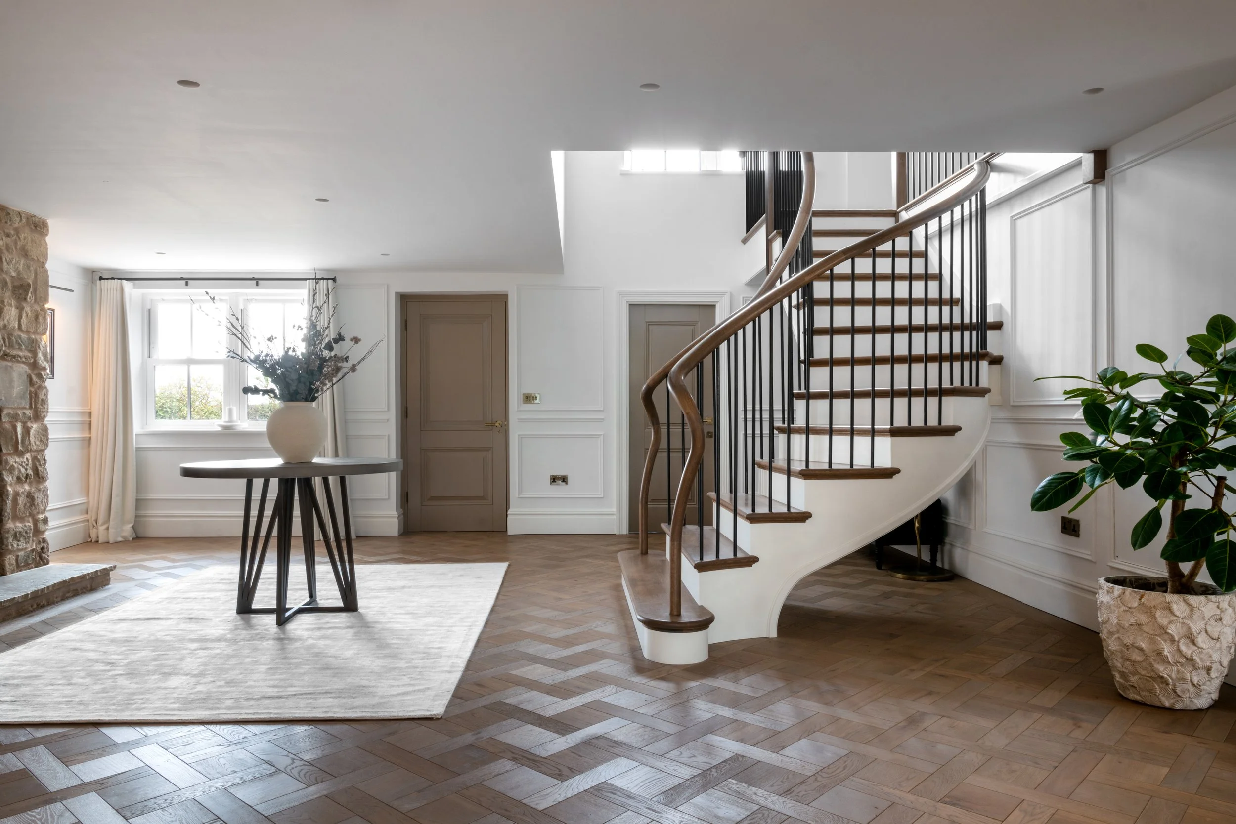 Interior view of a modern home with a wooden staircase, black metal railings, a table with a large beige vase filled with dried flowers, a floor lamp with a beige shade, and white paneled walls.