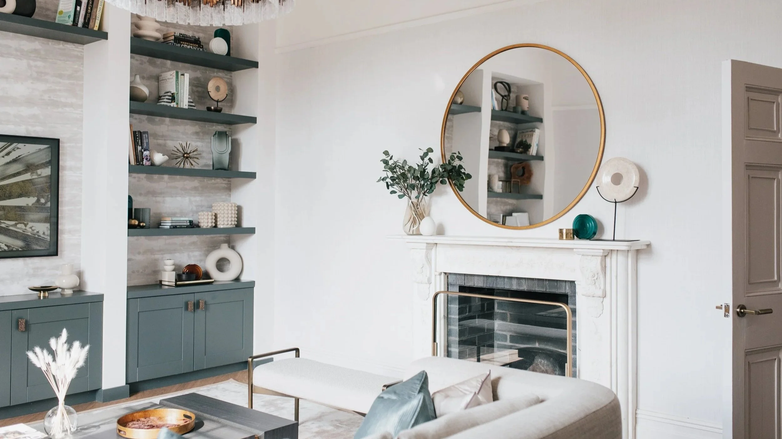 Living room with bespoke blue-green built-in shelving and cabinetry, styled open shelves, and a white fireplace with round brass-framed mirror