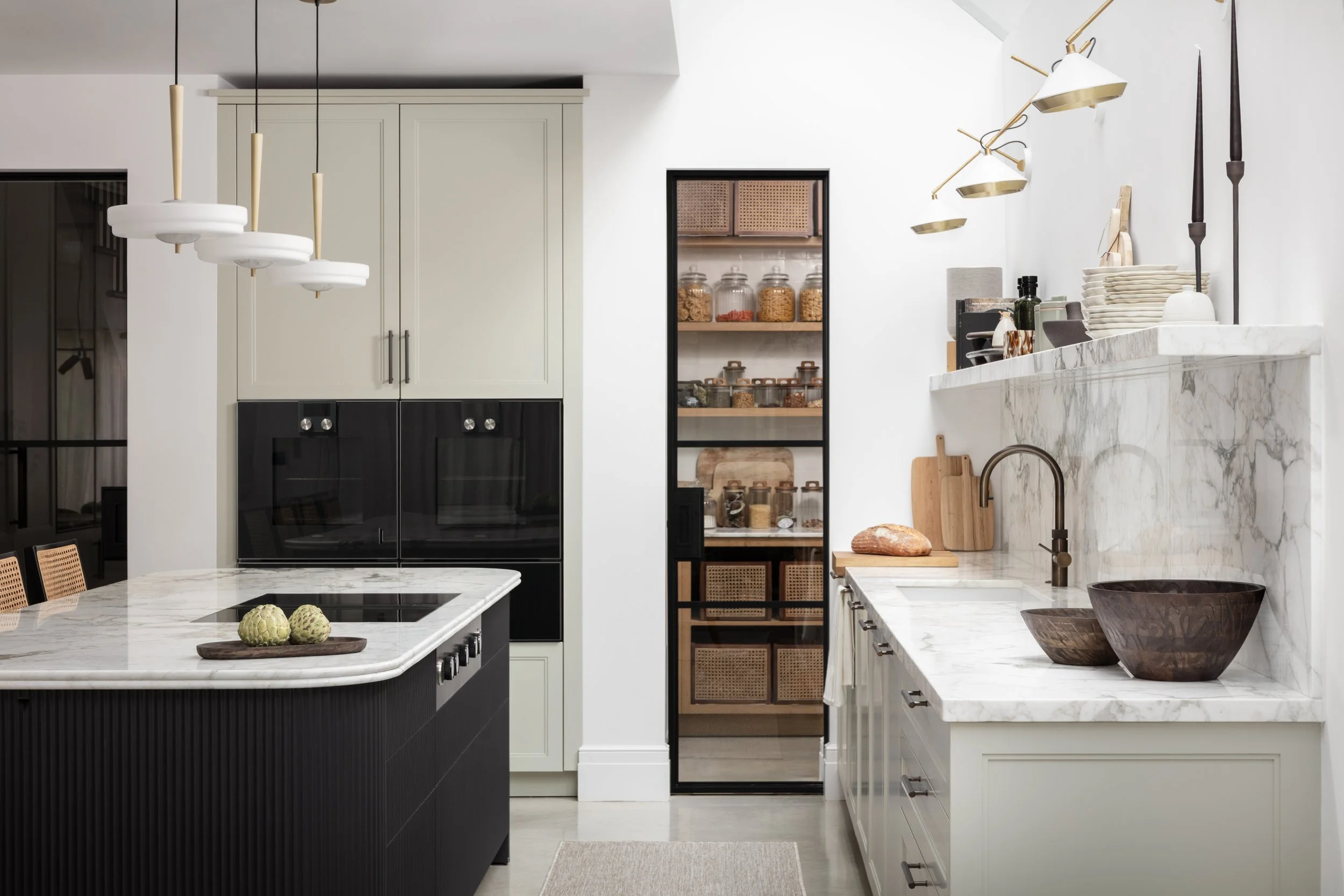 Modern kitchen with white marble countertops and a black kitchen island featuring three artichokes on a wooden tray. There is also a framed pantry with glass jars, beige cabinetry, and a marble backsplash. Hanging light fixtures and a window are visible.