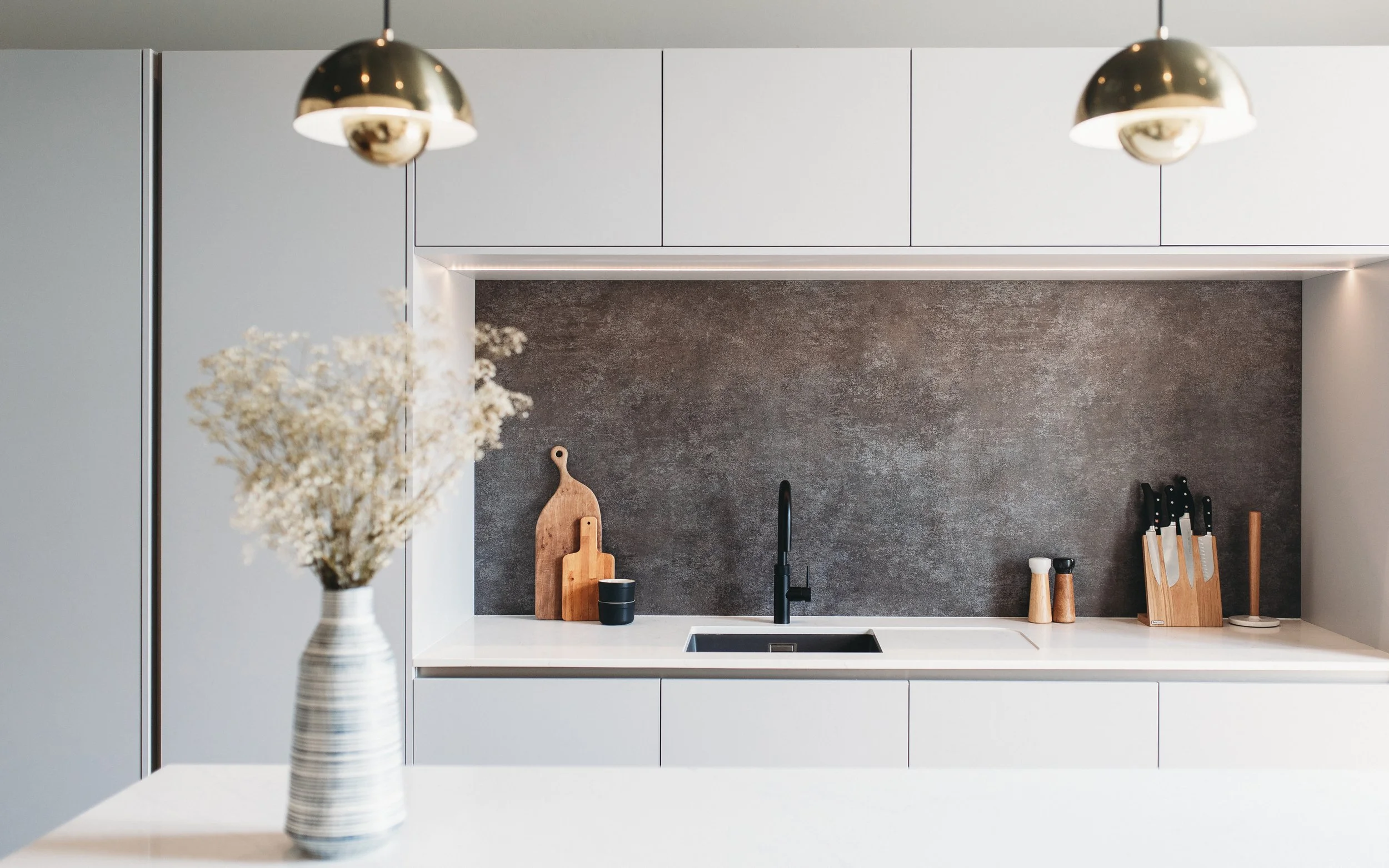 Minimalist kitchen with handleless white cabinetry, dark textured splashback, integrated lighting and black tap