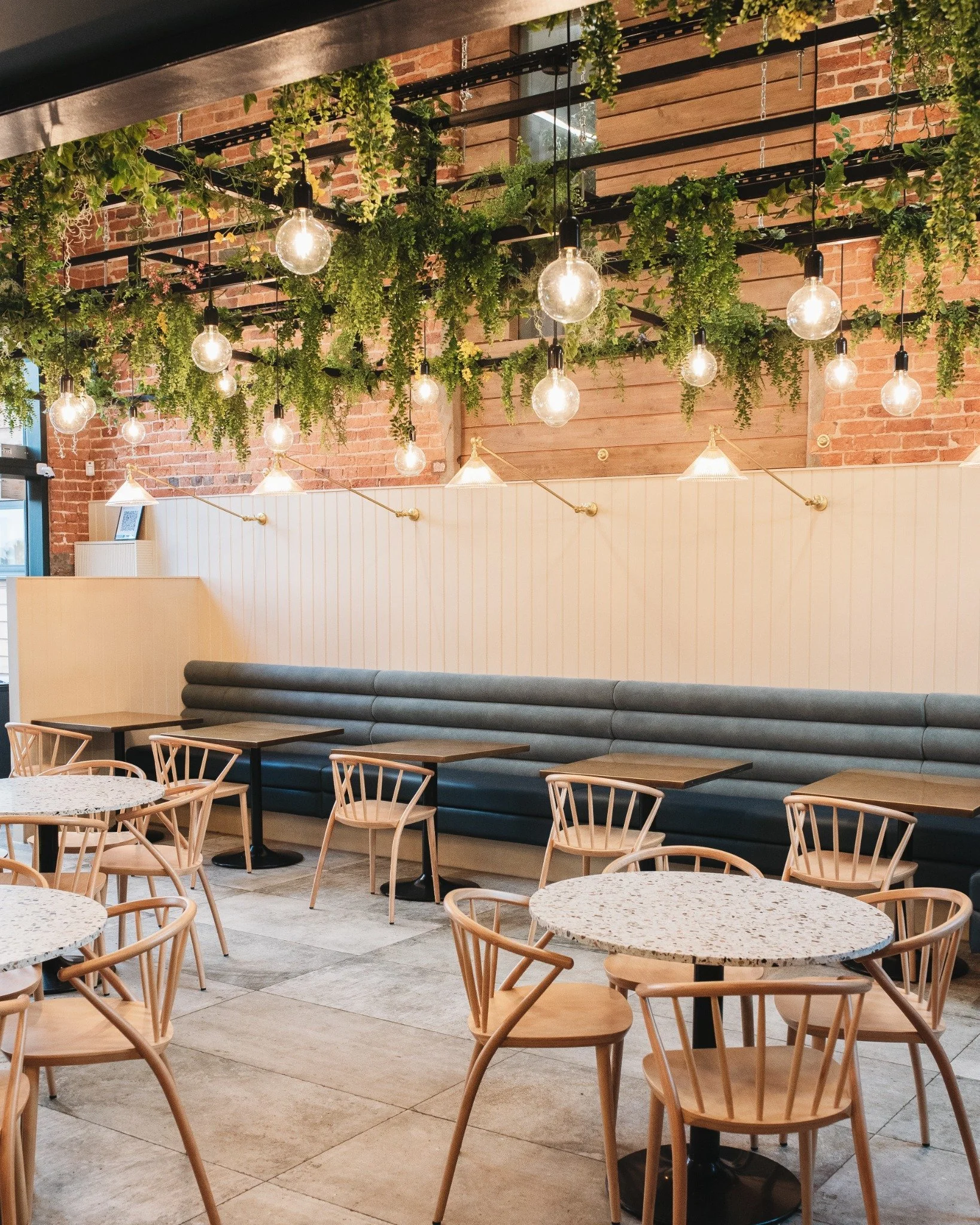 Restaurant interior with upholstered banquette seating, white timber wall panelling, brass wall lights and exposed brick with hanging greenery