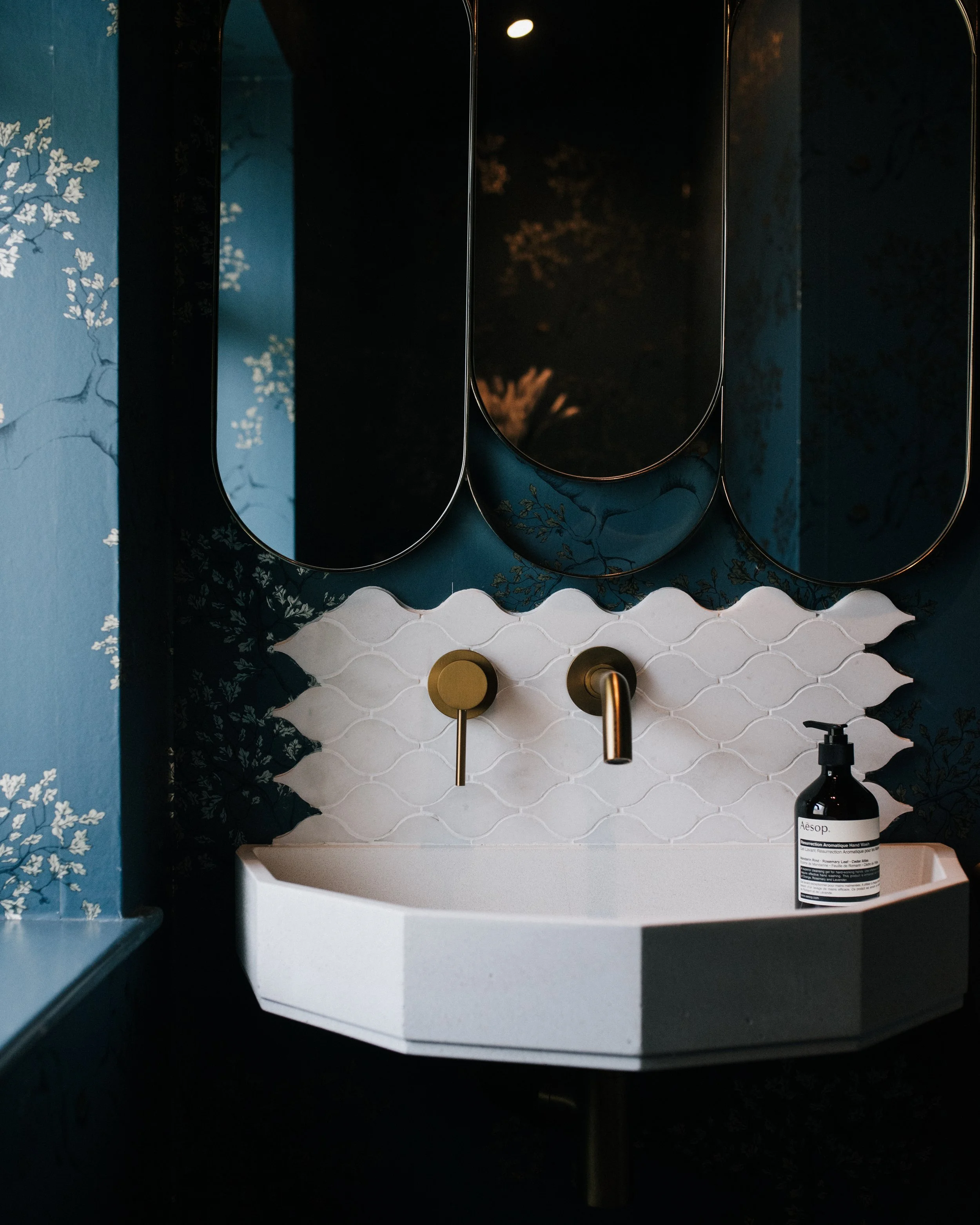 Cloakroom basin with scalloped white tiles, brass wall-mounted taps and decorative mirrors against patterned wallpaper
