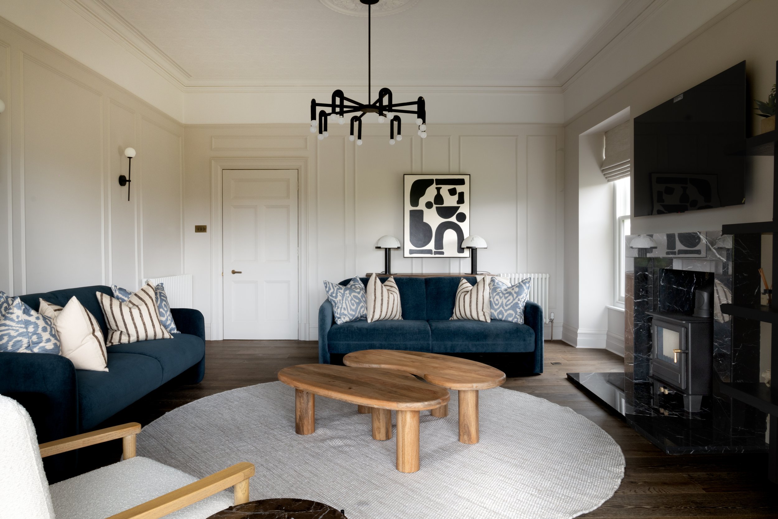 Living room with twin blue velvet sofas, layered patterned cushions, sculptural wooden coffee tables and panelled walls beneath a modern black chandelier