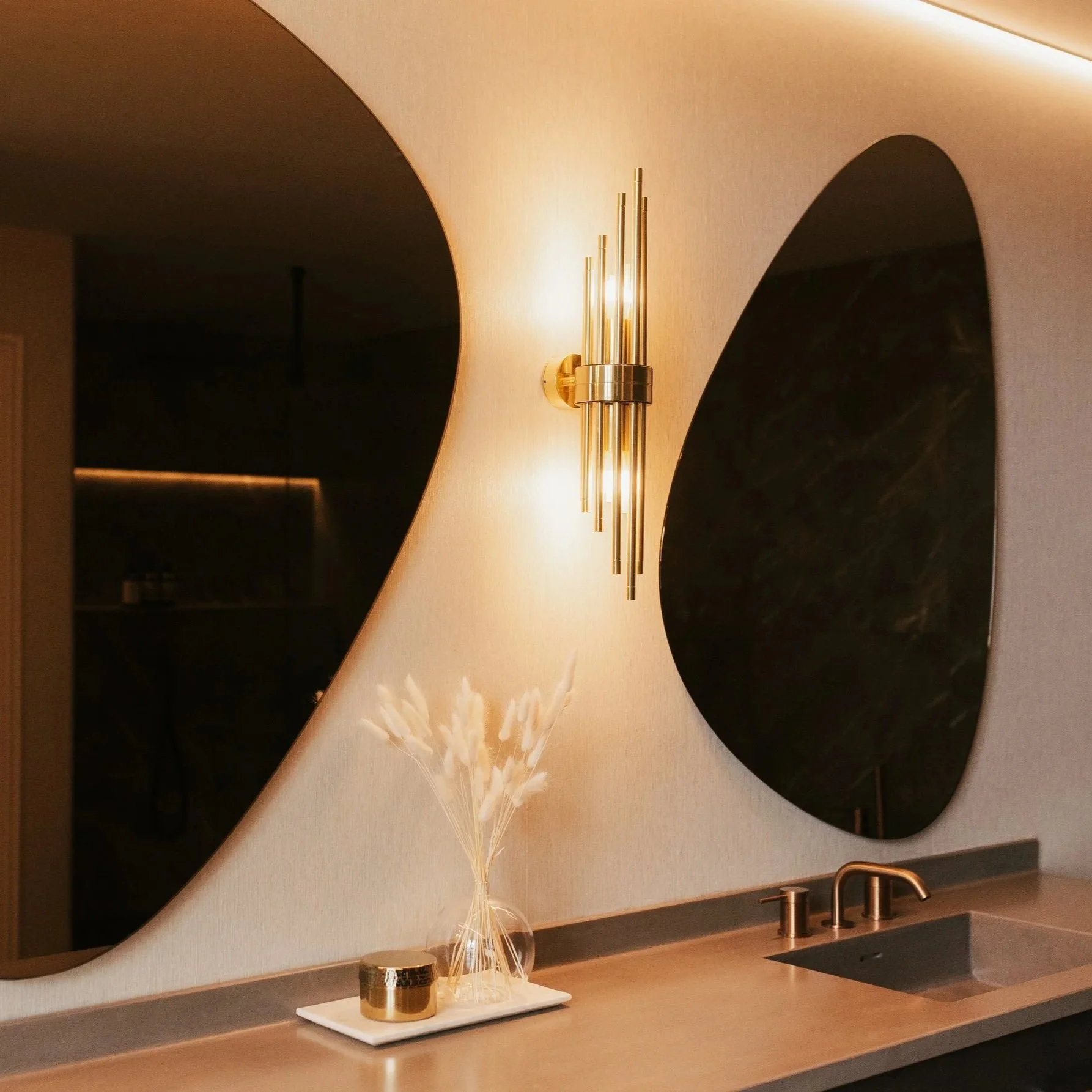 Bathroom vanity with sculptural oval mirrors, warm brass wall light and stone countertop with integrated sink