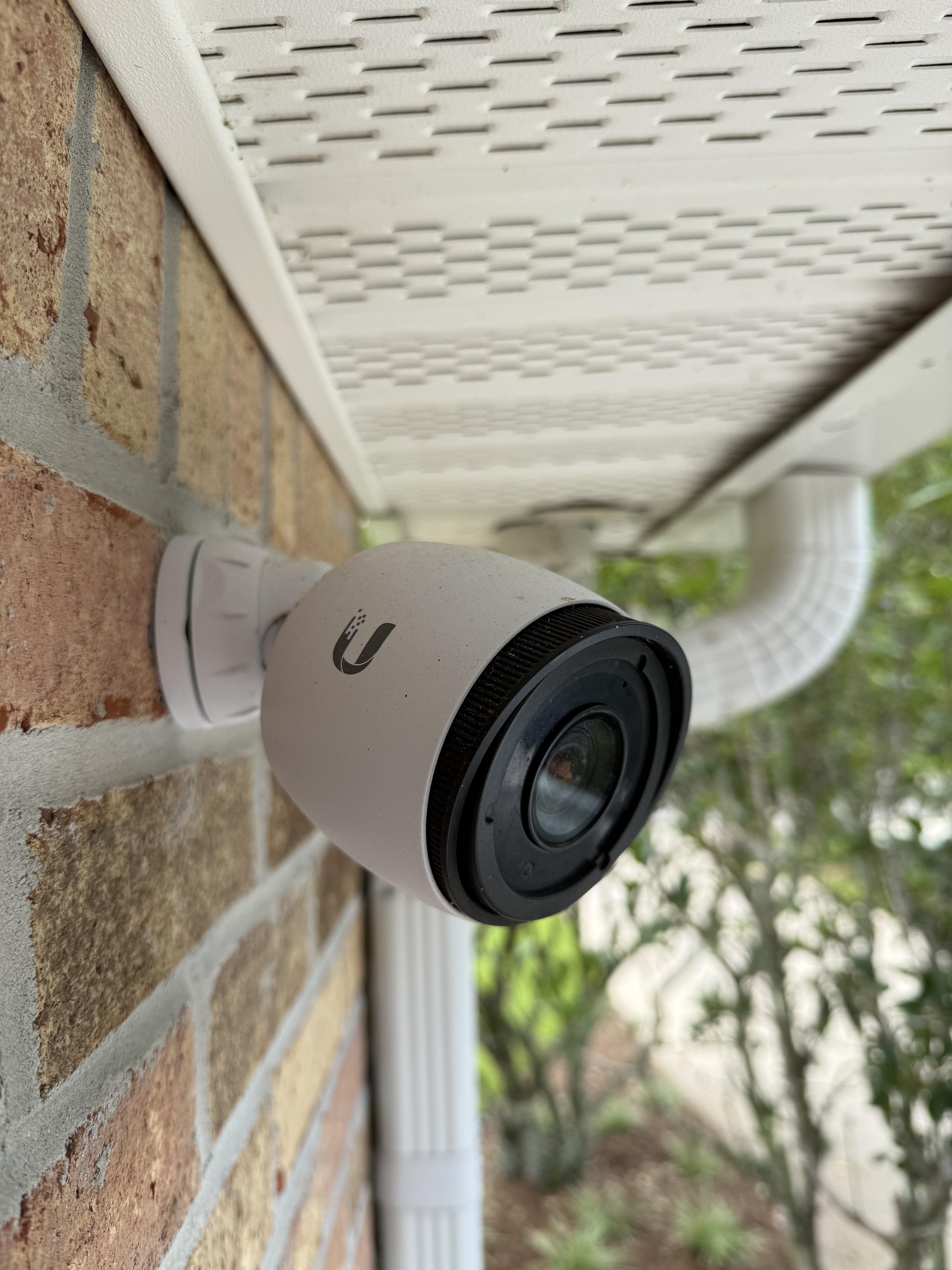 A white security camera mounted on a brick wall outside, near a white vent pipe, with greenery in the background.