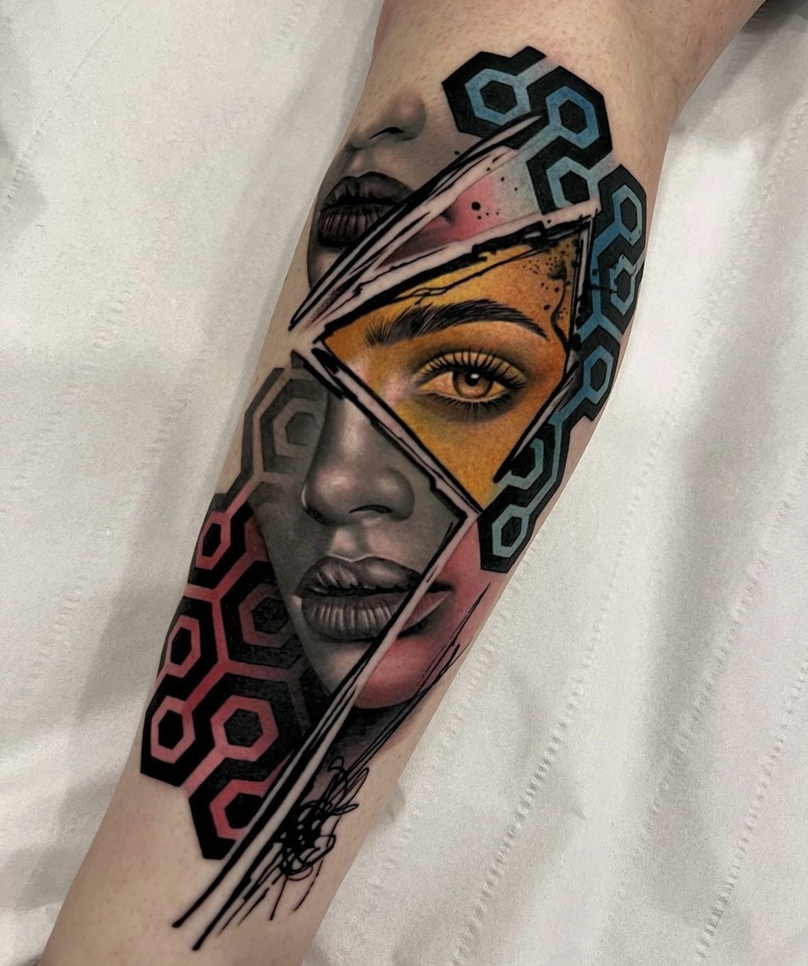 Tattoo of a woman’s face with geometric elements, including a yellow triangle around one eye and black, blue, and pink accents.