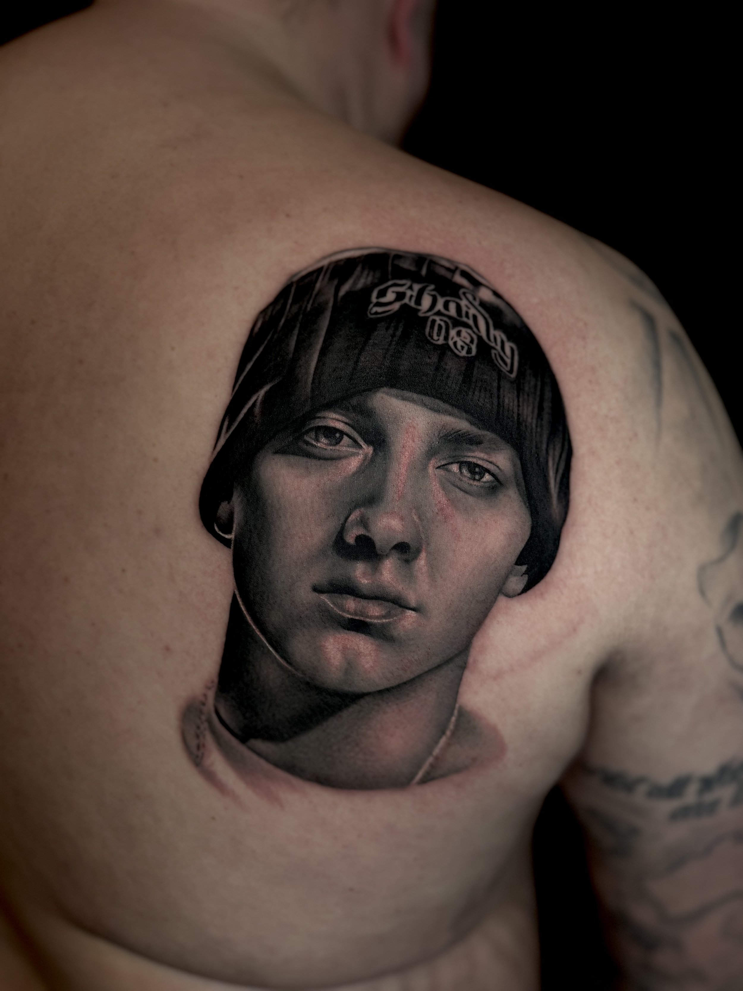 Tattoo of a young man's realistic face on person's upper chest, wearing a black beanie.