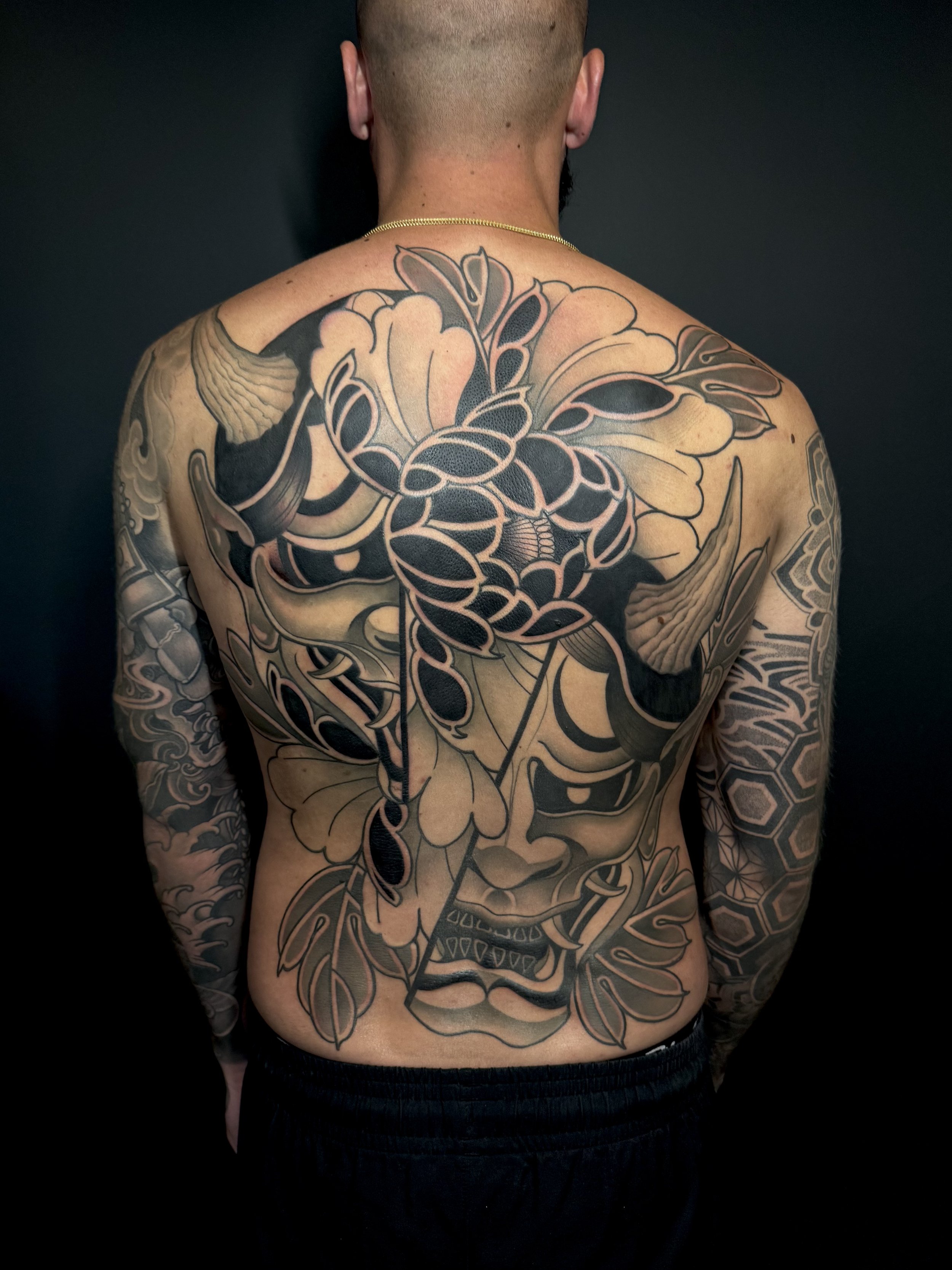 A man's back with a large Japanese-style tattoo featuring a fierce face, flowers, a snake, and intricate geometric designs in black and beige ink.