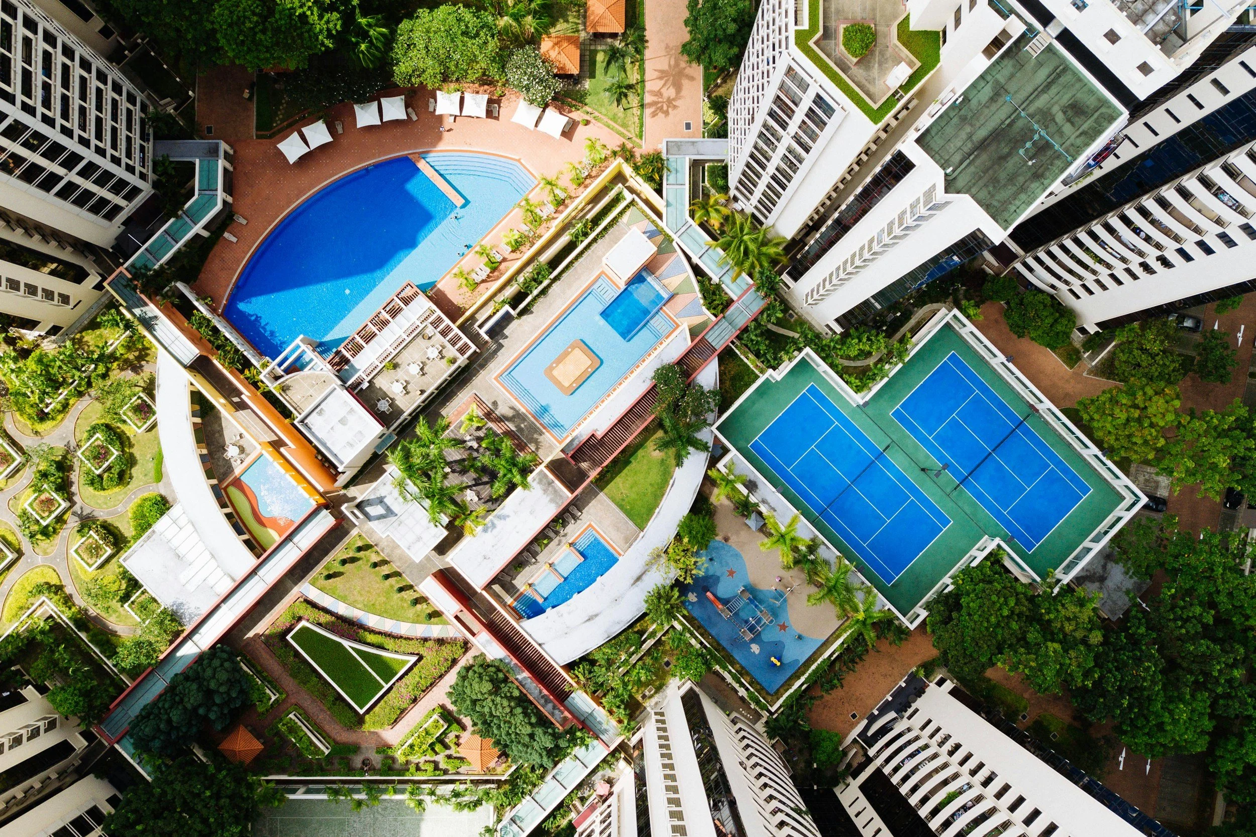 Aerial view of a high-rise residential complex featuring multiple swimming pools, tennis courts, courtyards, and landscaped gardens surrounded by tall buildings.