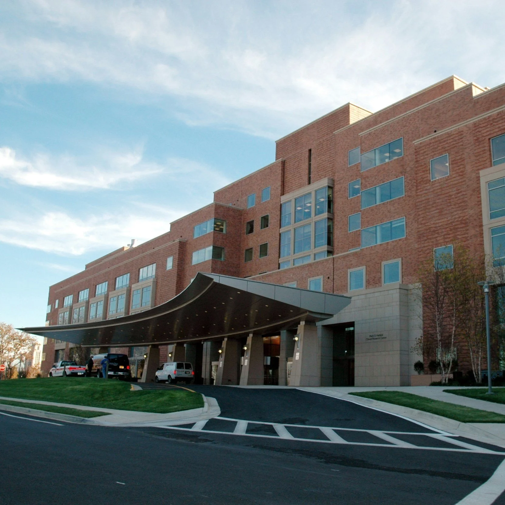 Modern brick building with large windows and a curved entrance canopy, vehicle lane and landscaped greenery in the foreground.