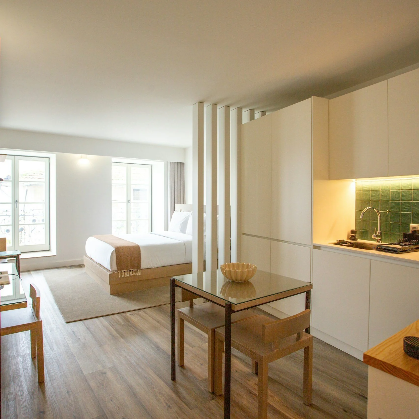 Modern studio apartment with a bed near windows, small dining table, kitchenette with green tiled backsplash, and wooden flooring.