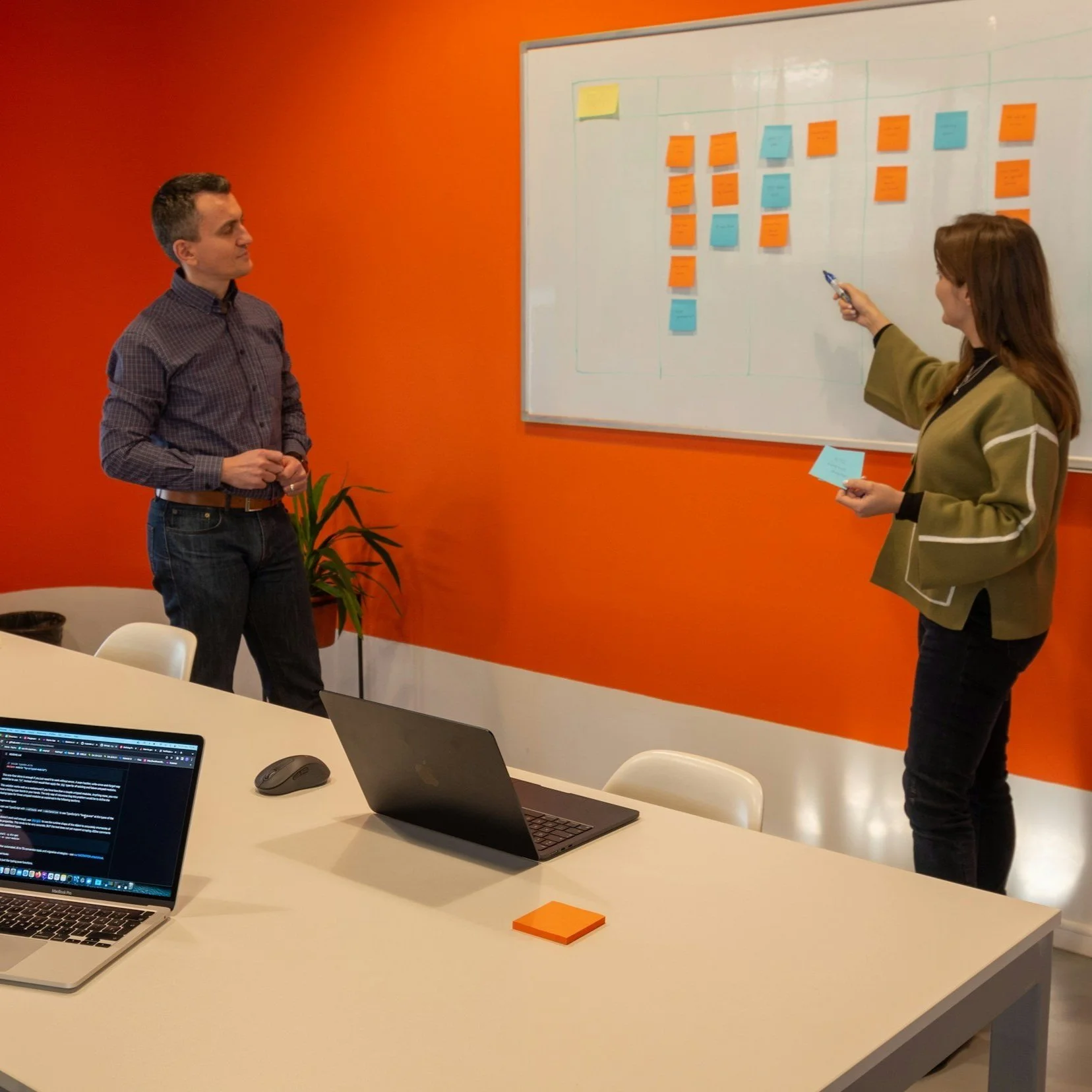 Two people are conducting a meeting in an office with a whiteboard and sticky notes. A man stands near the left side of the image, wearing a checkered shirt and jeans. A woman stands near the whiteboard, holding a blue sticky note and a marker. The whiteboard has several notes on it, with some yellow, orange, and blue notes pinned or attached to it. There are laptops and sticky notes on the white conference table in the foreground.