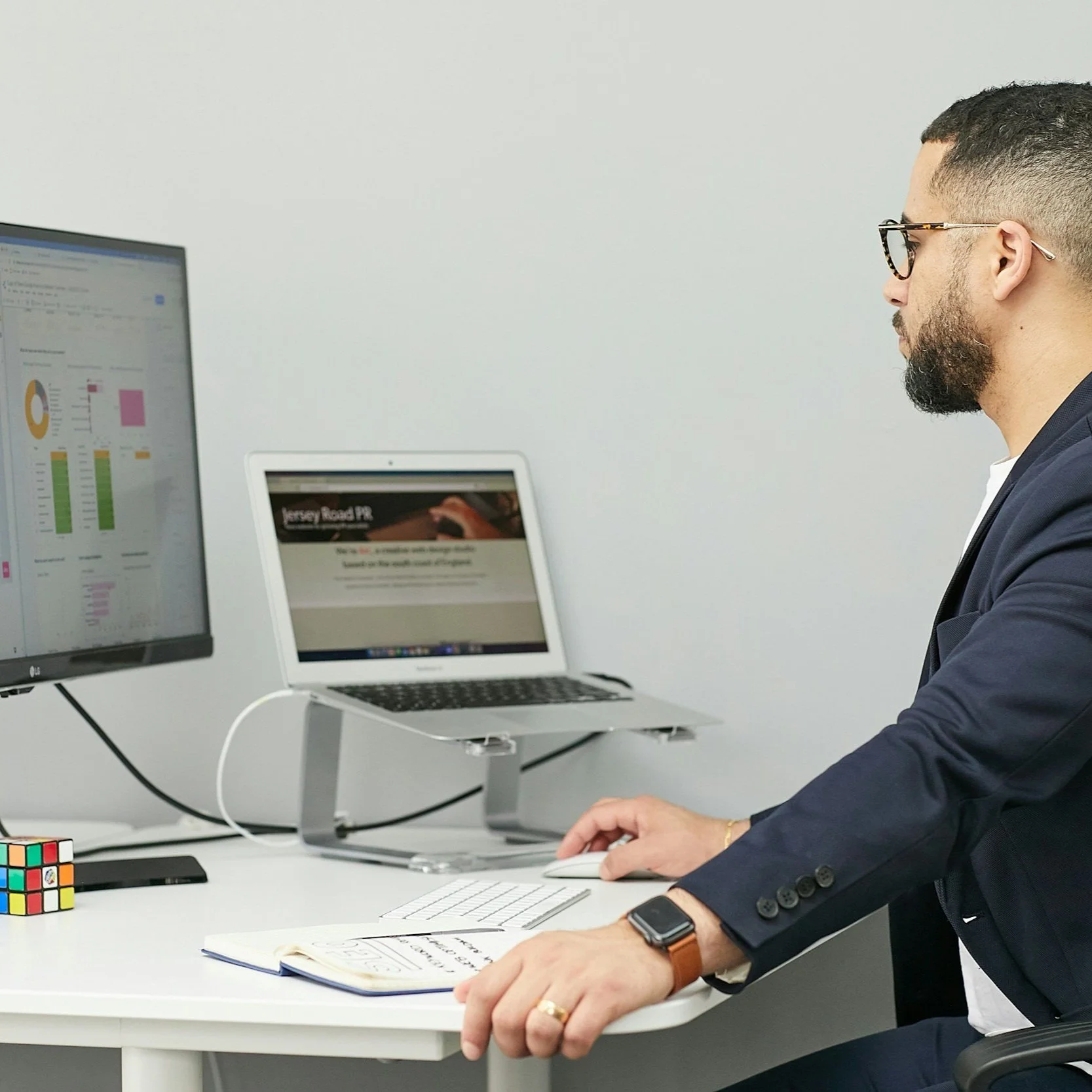 Man wearing glasses and a suit working at a desk with two monitors, a laptop, and a notebook.