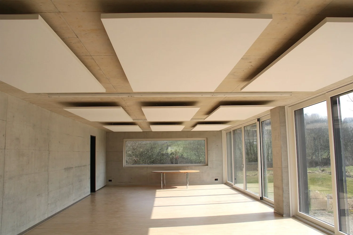 Heller Raum mit Sichtbeton und bodentiefen Fenstern, an der Decke quadratische weisse Akustik-Deckensegel