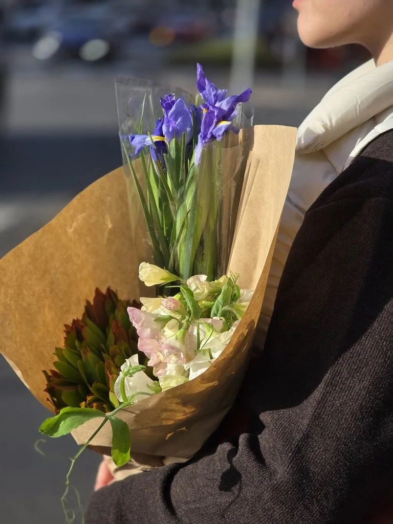 A person holding a bouquet of colorful flowers wrapped in brown paper, with purple, white, and green flowers visible.