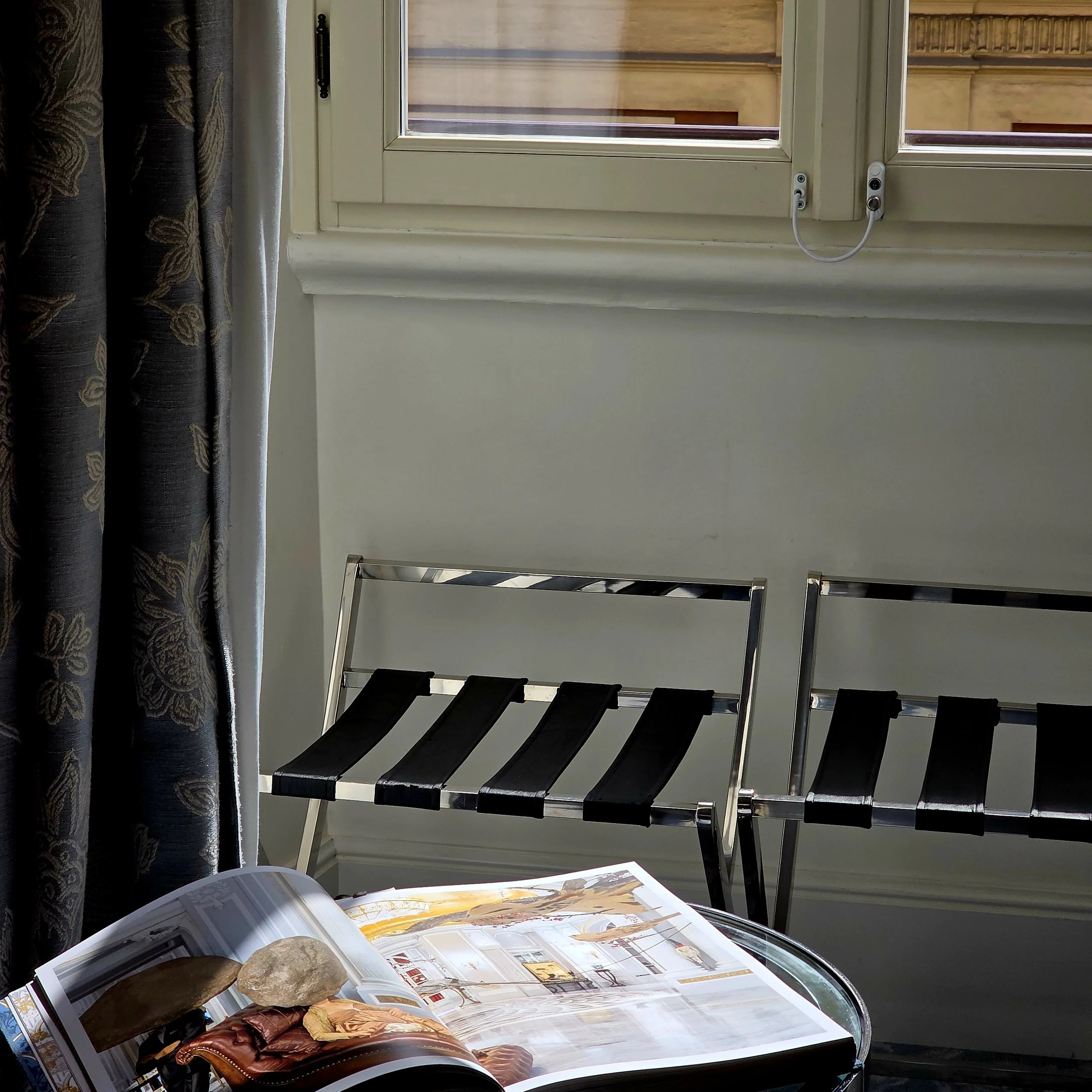 Interior scene with a glass table holding an open magazine, two black and chrome chairs with strap seating, a window with partly open blinds, a curtain on the left, and a white wall.