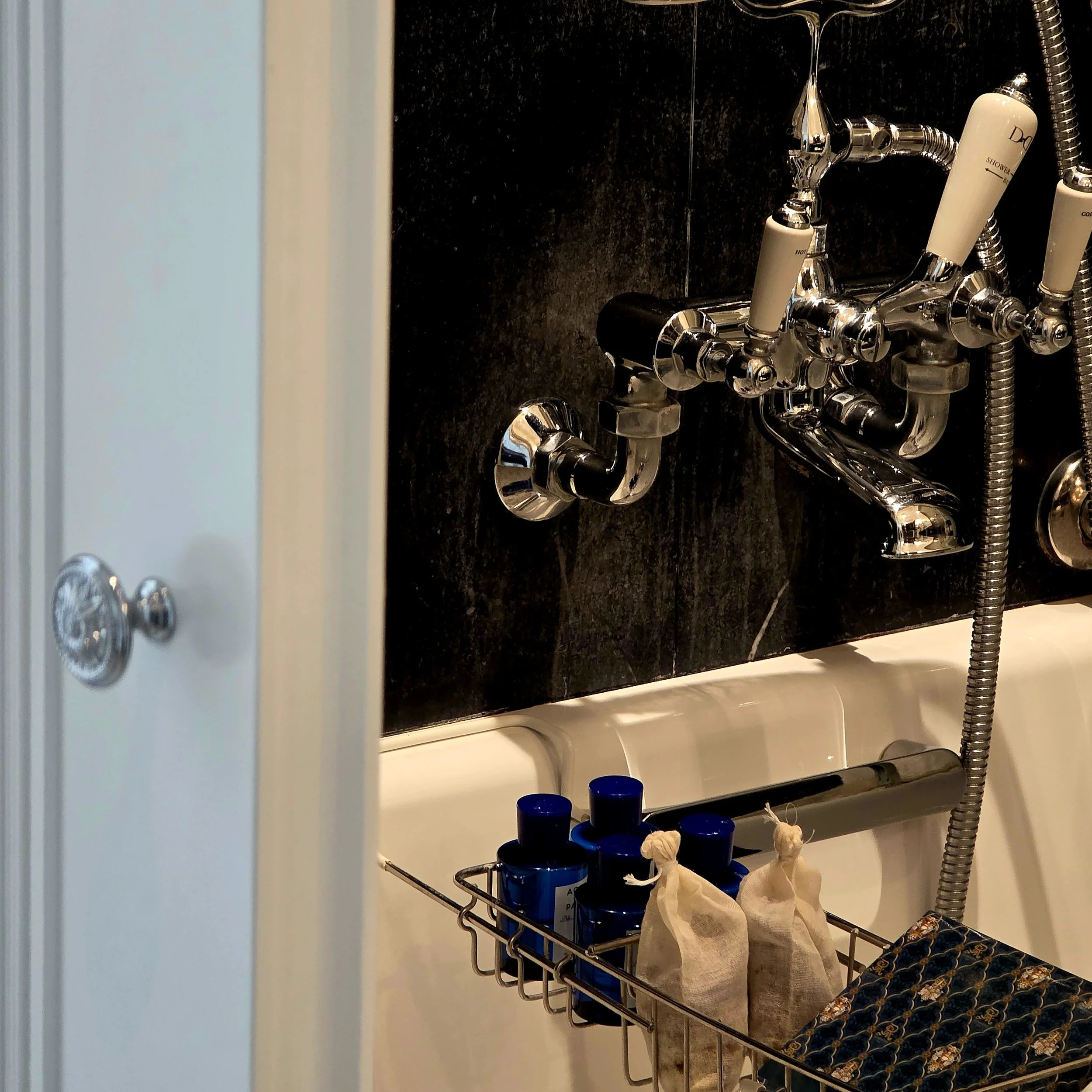 Bathroom sink with a black tiled wall, silver faucet, and shower controls, with blue bottles and small cloth bags in a metal rack above the bathtub.