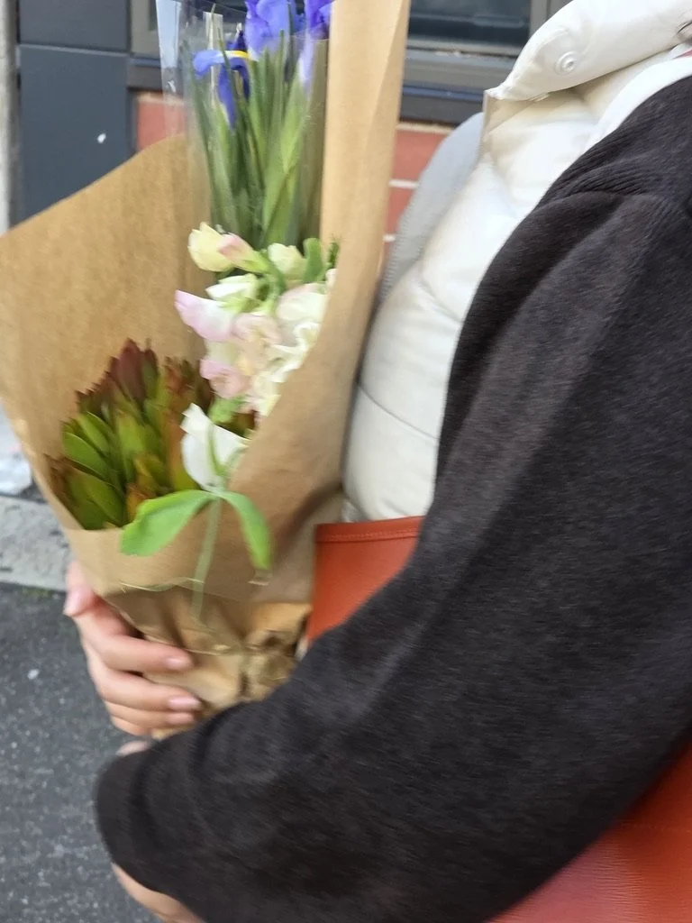 Person holding a bouquet of purple, white, and pink flowers wrapped in brown paper, with a background of a brick and wooden wall.
