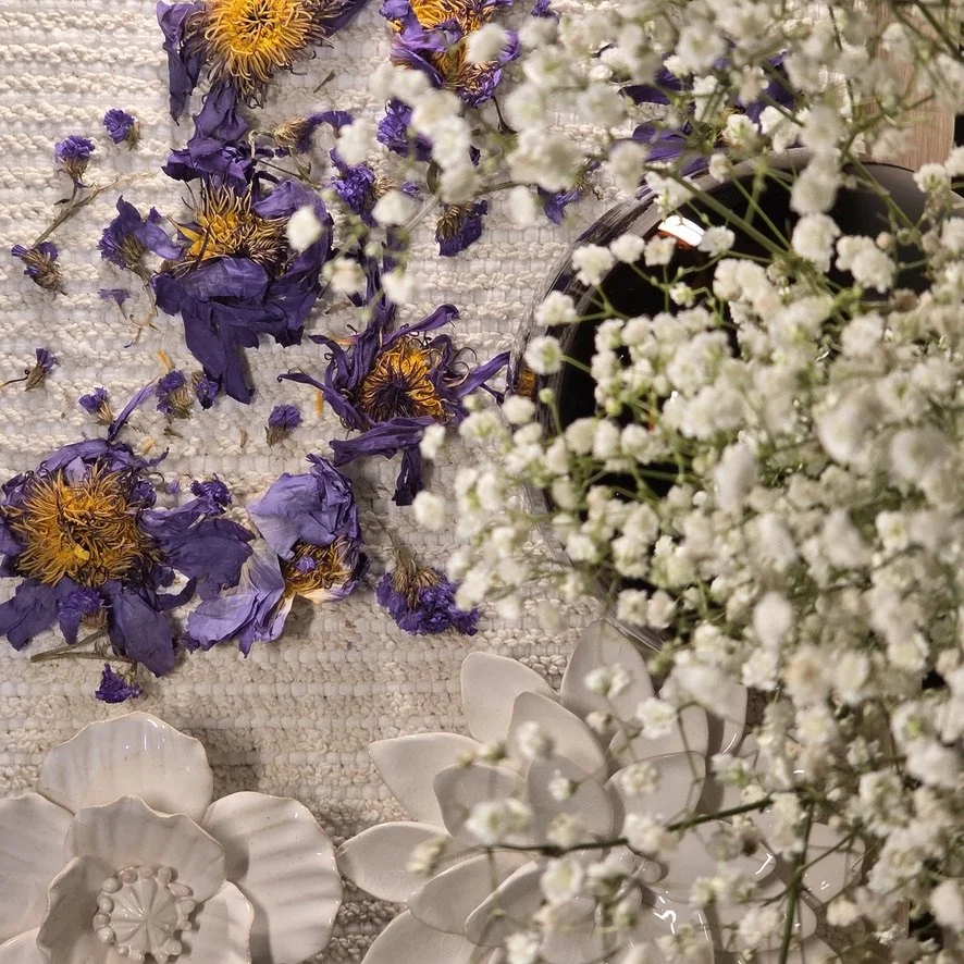 A flat lay of dried purple and yellow flowers, small white baby's breath flowers in a vase, and decorative ceramic flower dishes on a textured light-colored surface.