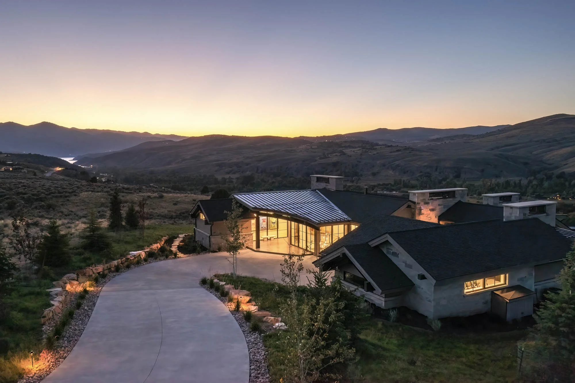 A modern custom home in Park City with large glass windows and a metal roof is illuminated at dusk, situated on a hillside with a winding driveway and surrounded by natural landscape of rolling hills and mountains in the background.