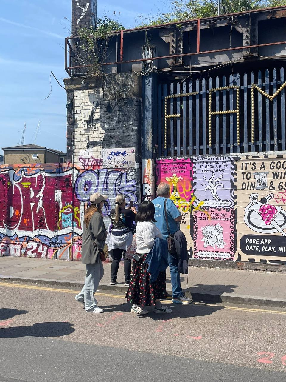 A group of five people standing on a sidewalk in front of a graffiti-covered wall with pink posters. One of the posters says 'It's a good day to book a dick appointment.' The people are looking at the posters and graffiti, with some holding bags. The