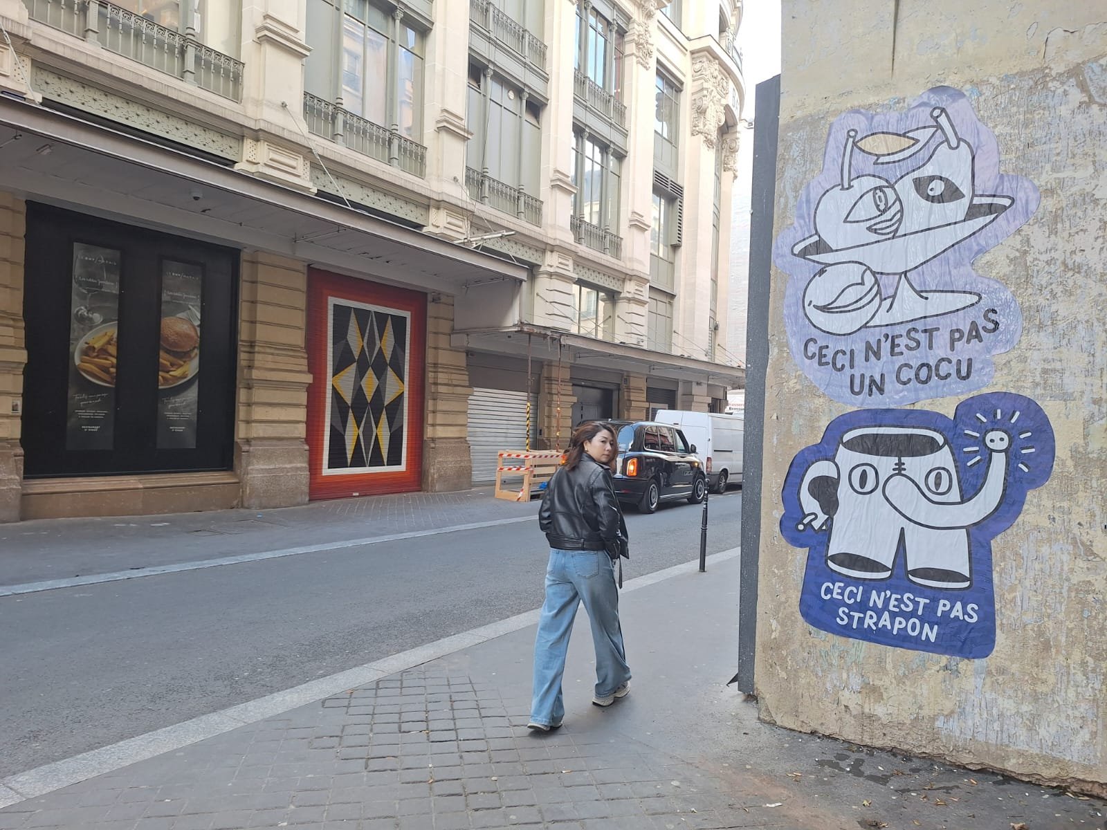 A woman with long hair, wearing a black leather jacket and light blue jeans, walking on a city sidewalk. Next to her, on a wall, are two stickers: one showing a cartoon fish with the French text 'Ceci n'est pas un cocu' and a second showing a cartoon