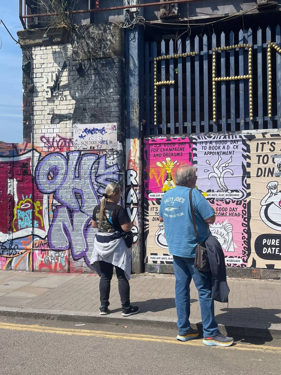 Two people stand on the sidewalk outside a colorful street wall with graffiti and posters. A woman with a ponytail and black outfit and a man with gray hair in a blue T-shirt are facing the wall, looking at posters that contain text related to dating