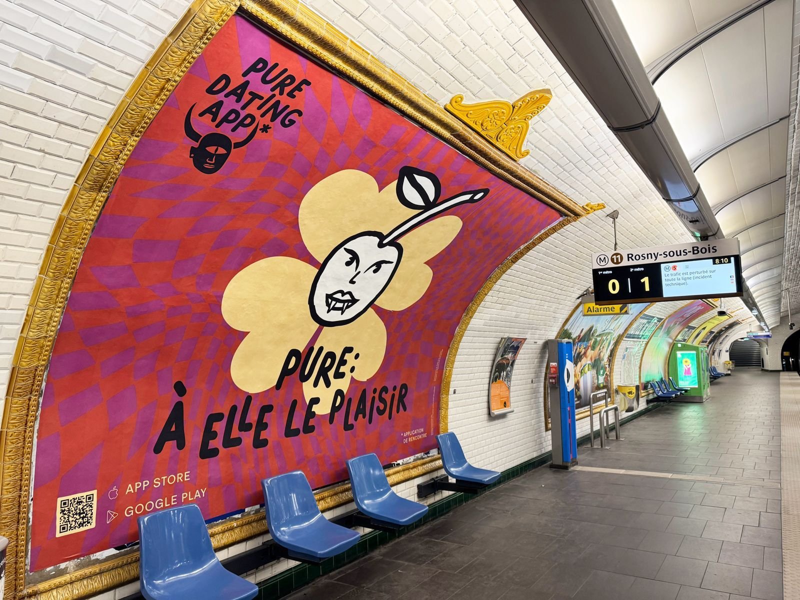 Empty subway platform with colorful advertisement featuring a cartoon pear with a human face and text in French, with train schedule display showing train to Rosny-sous-Bois.