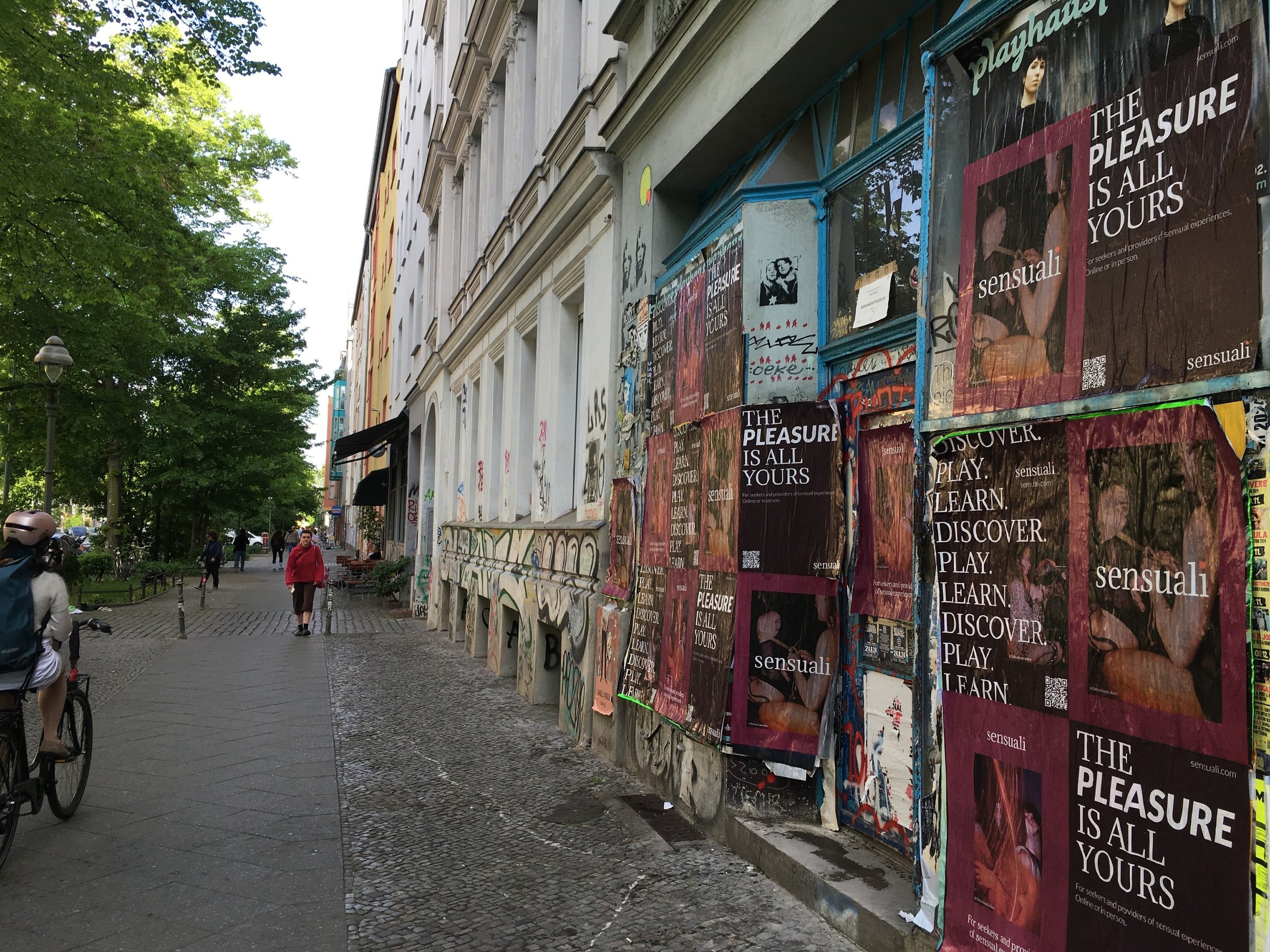Street scene with a sidewalk, people walking and riding bicycles, and a building covered in posters including ads for Sensuali with the slogan 'The pleasure is all yours,' and graffiti on the wall.
