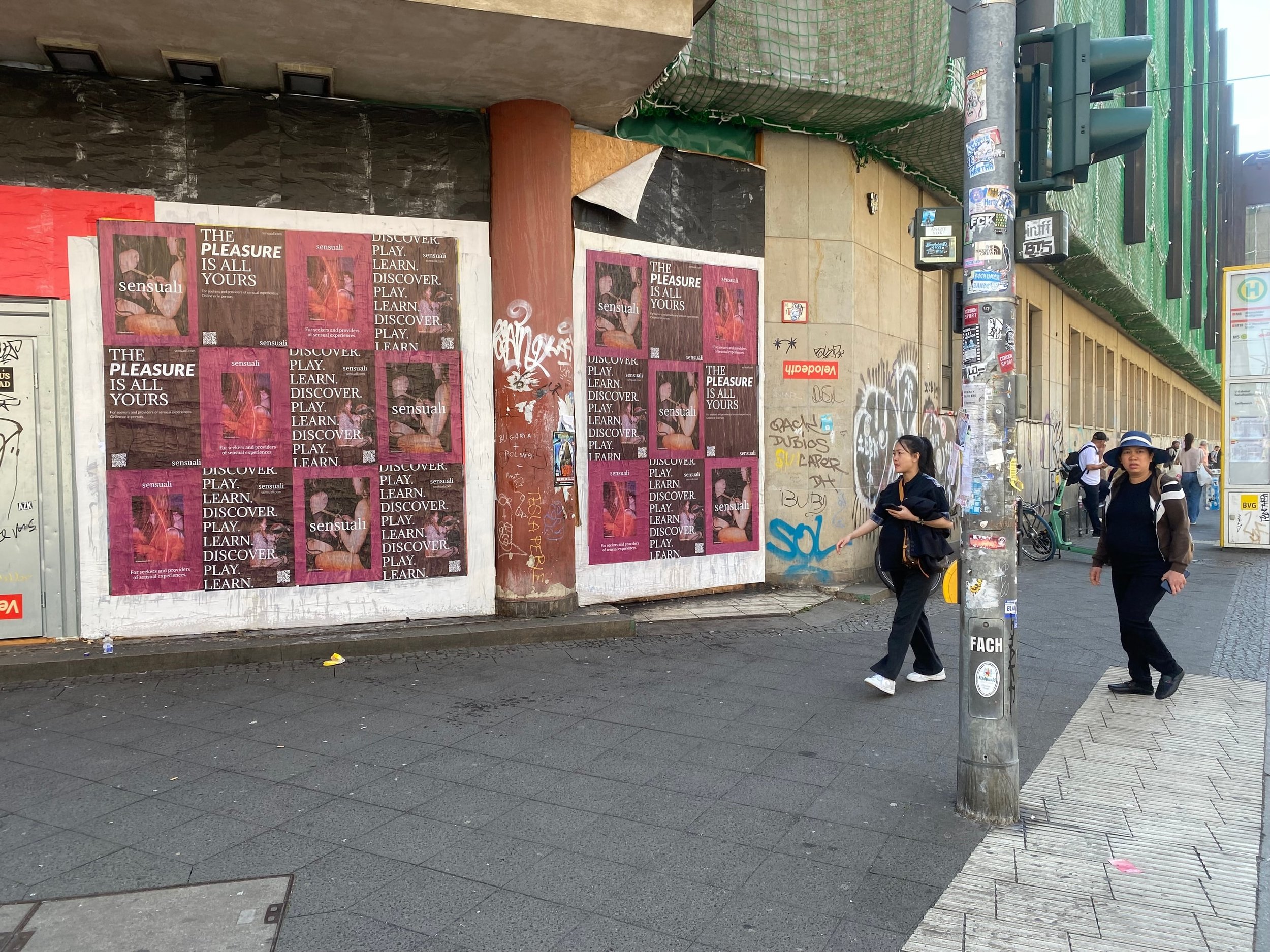 Street scene with graffiti-covered wall, posters advertising βSensual,β pedestrians crossing, and a lamppost covered in stickers and graffiti.
