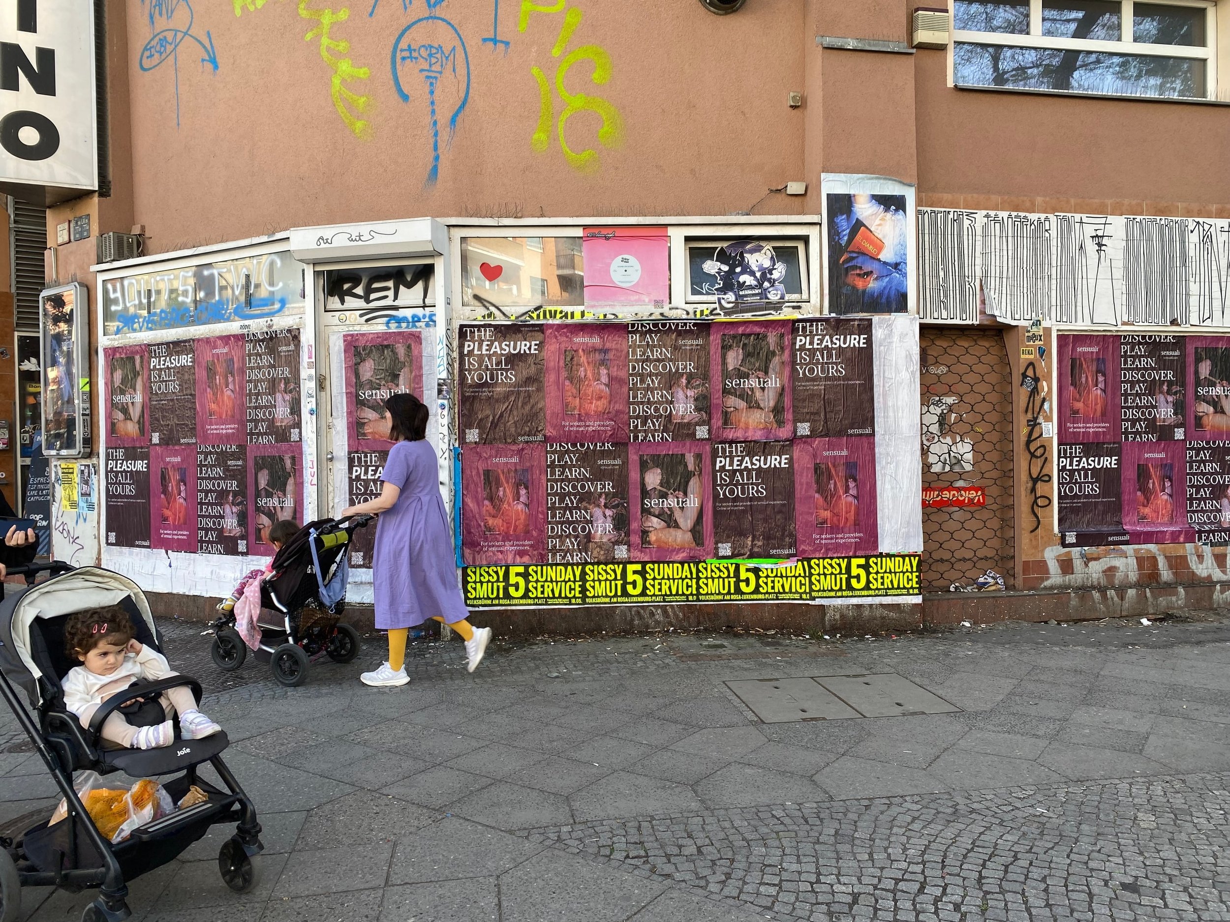 Street scene with a woman and two children, one in a stroller, in front of a building covered with posters, graffiti, and advertisements.