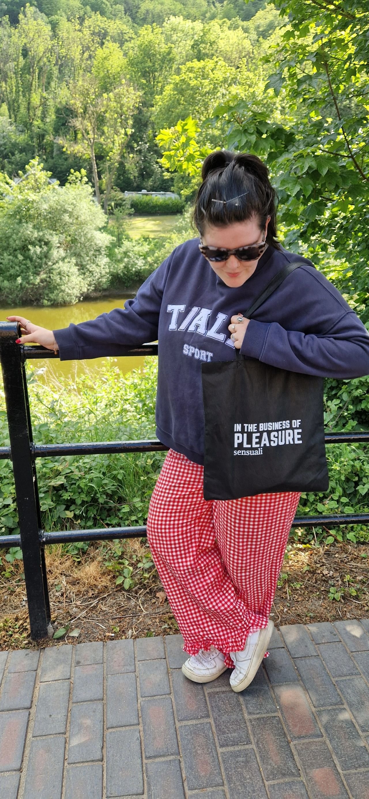 A woman standing outdoors near a river, wearing sunglasses, a navy sweatshirt, red gingham pants, and white sneakers, holding a black tote bag with white text.