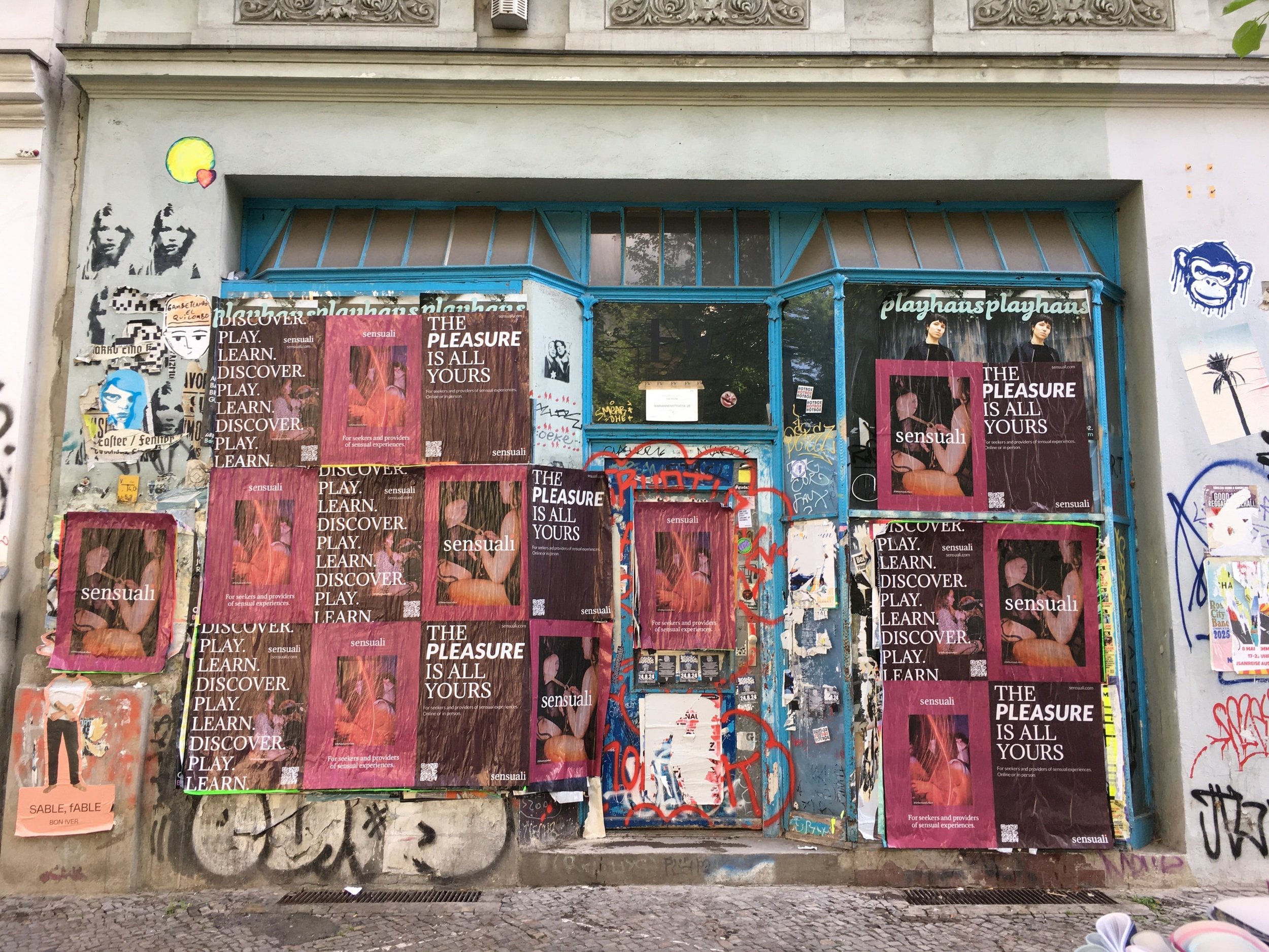 Graffiti-covered wall with blue metal window frame, covered in multiple posters advertising sensations, pleasure, discovery, and sensual experiences, some with images of women and text like 'The pleasure is all yours' and 'Sensual'. There is also gra