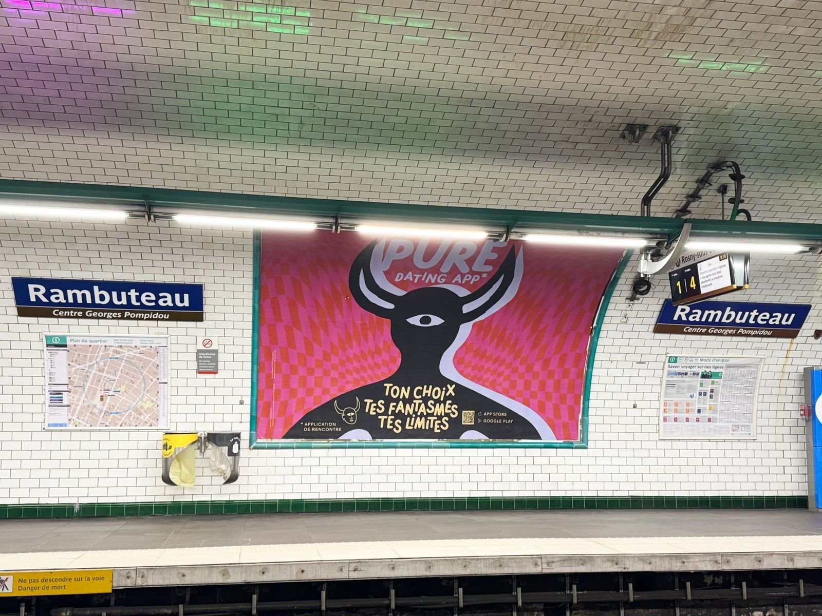 Subway station platform at Rambuteau with a large pink and black advertisement featuring a stylized creature with horns and a third eye, and surrounding signs and maps.