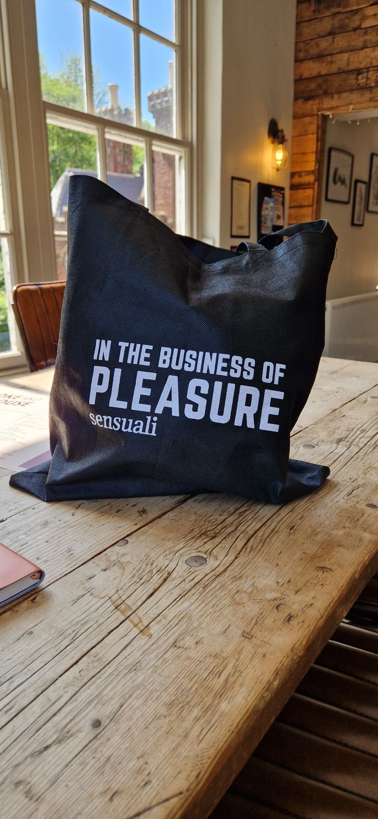 Black tote bag with the text "In the business of pleasure sensuali" placed on a rustic wooden table in a bright room with large windows and framed pictures on the wall.