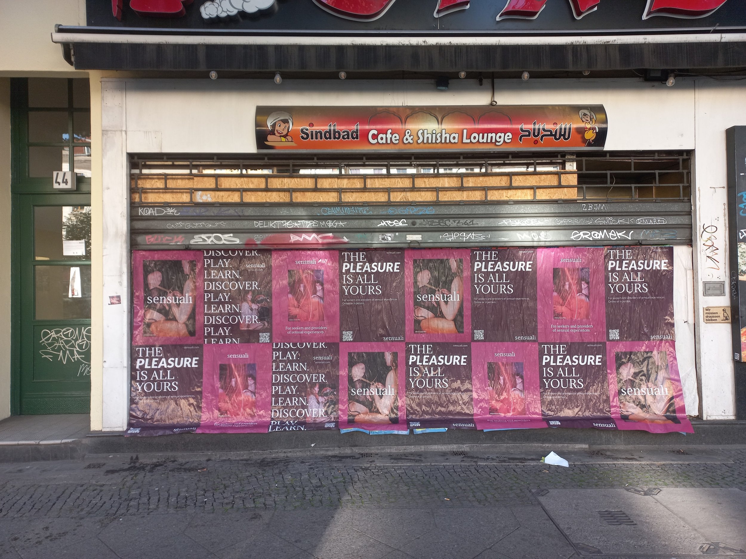 Closed storefront with multiple pink and black posters advertising sensual pleasure and discovery, overlapping a graffiti-covered metal shutter, with a sign above reading 'Sindbad Cafe & Shisha Lounge'.
