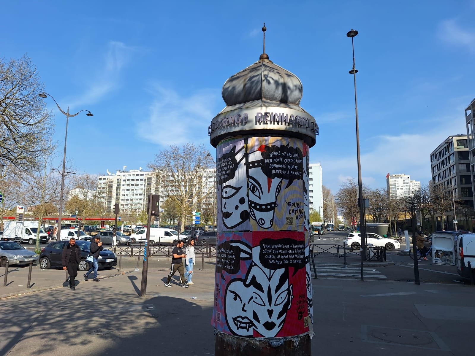 A city street scene with a decorated advertising column featuring black and white cartoon-style artwork and text, surrounded by pedestrians, parked cars, leafless trees, and modern buildings under a blue sky.