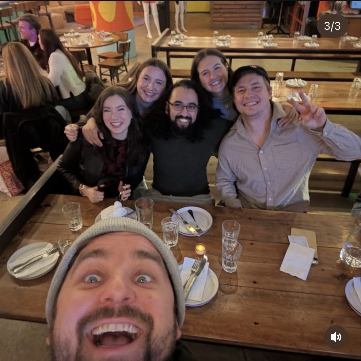 A group of six friends smiling and posing for a selfie at a restaurant table, with a man in a gray beanie in the foreground taking the photo. The table has plates, glasses, and utensils, and the background shows other diners and restaurant decor.