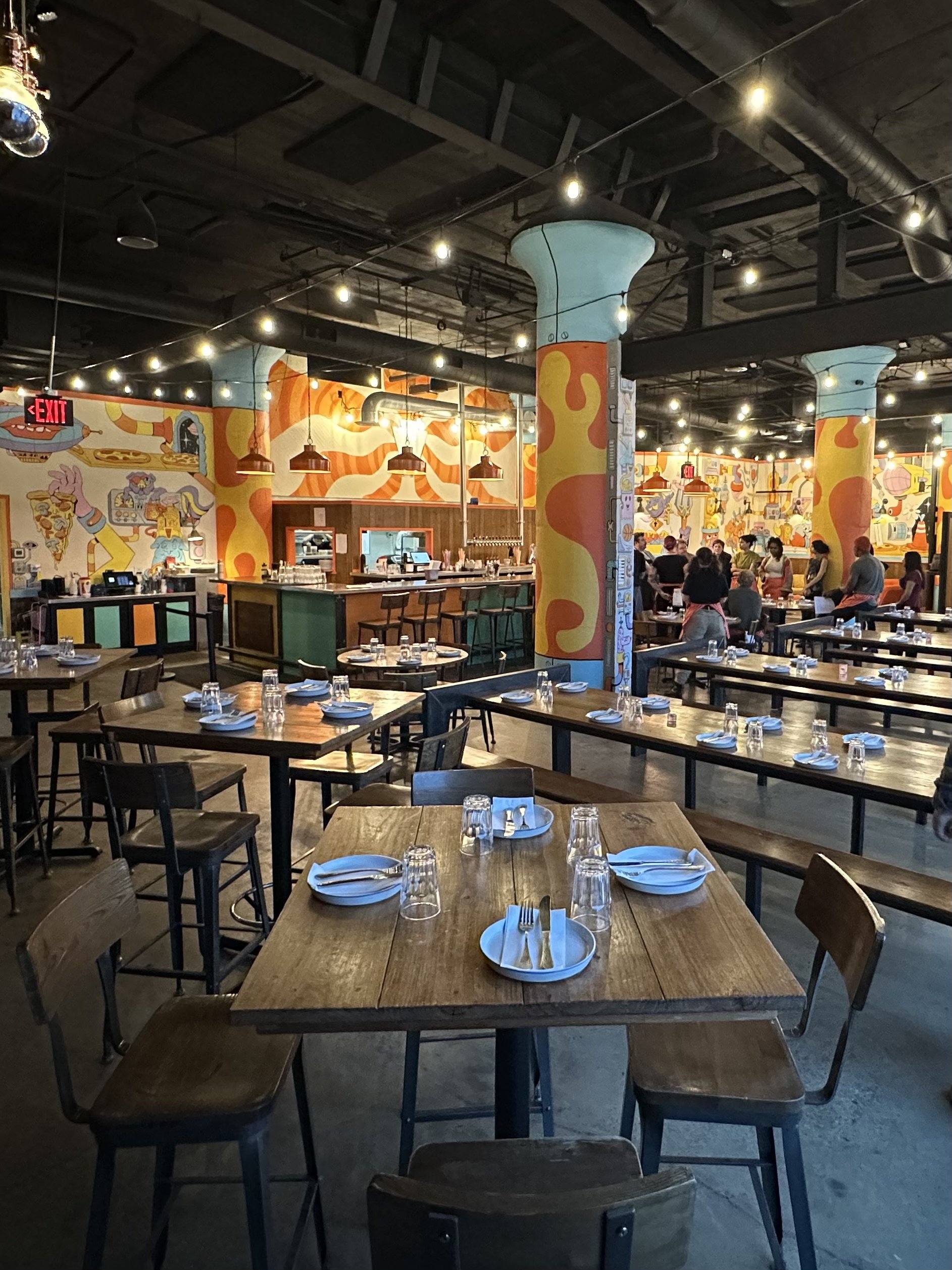 Interior view of a lively restaurant with wooden tables set with plates, glasses, and utensils, colorful graffiti-style murals on the walls, ceiling lights, and a bar area in the background with patrons and staff.