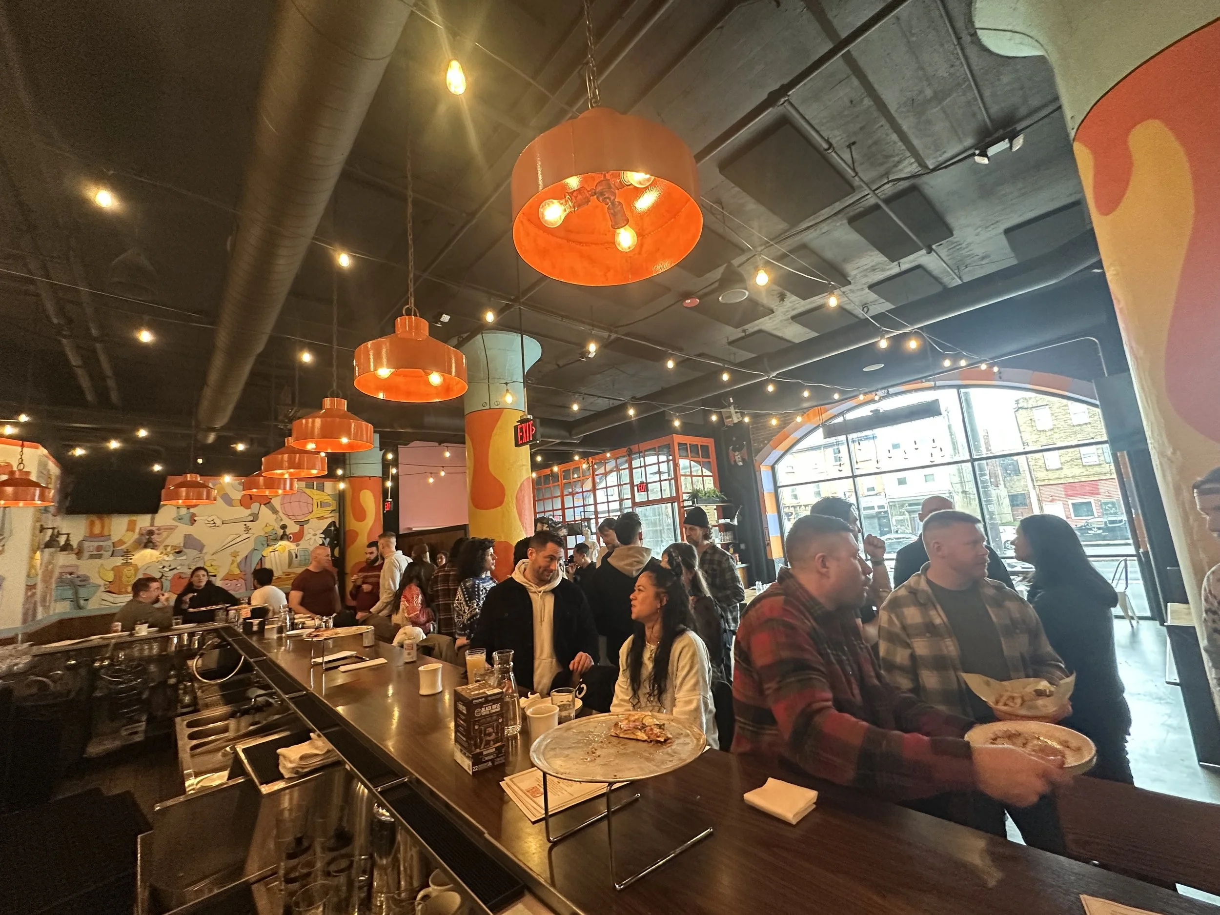 People inside a lively restaurant with colorful murals, orange lighting fixtures, and a large window at the front, some standing and chatting while others sit at the bar and tables.