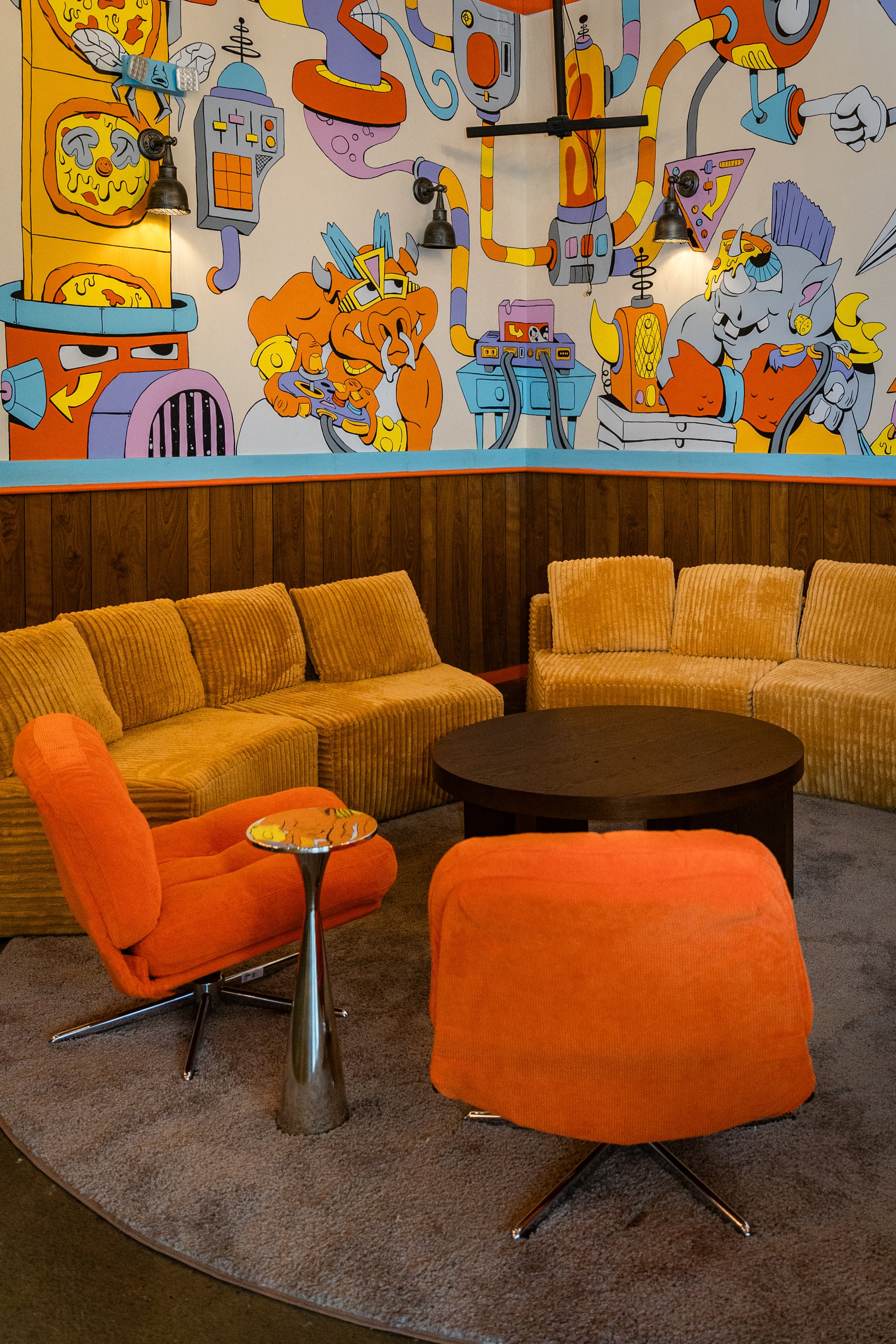 Our small group plushy lounge area with golden yellow sofas, orange chairs, and a round black coffee table. The wall features a vibrant, cartoon-style mural depicting whimsical creatures and abstract machines.